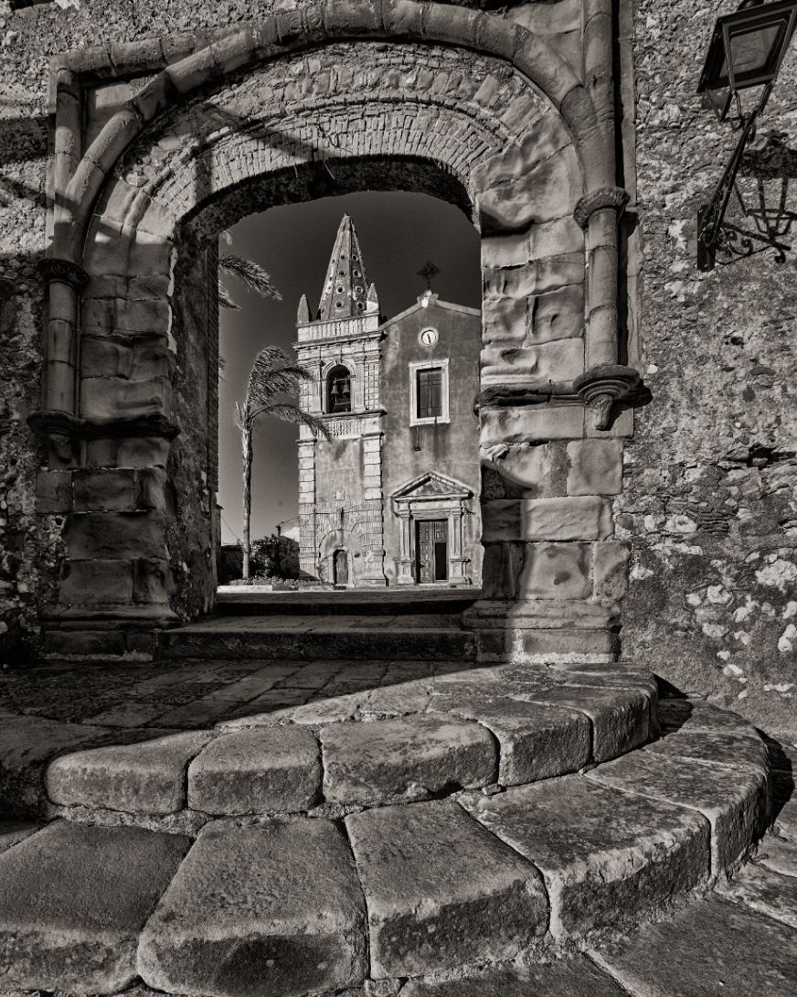 Black and white view through stone archway of historic church with bell tower in Sicilian village. Horvath-Photography-Sam-Sciarrino