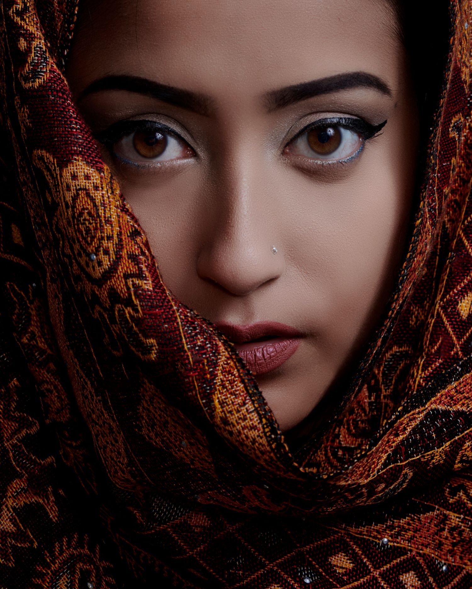 Close-up portrait with dramatic lighting shows a person wearing an ornate copper and gold patterned head covering.