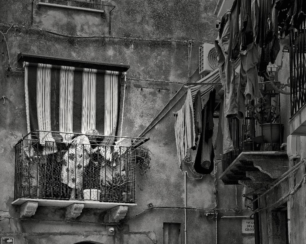 A black and white photograph shows clotheslines with laundry hanging between old apartment buildings in Italy.