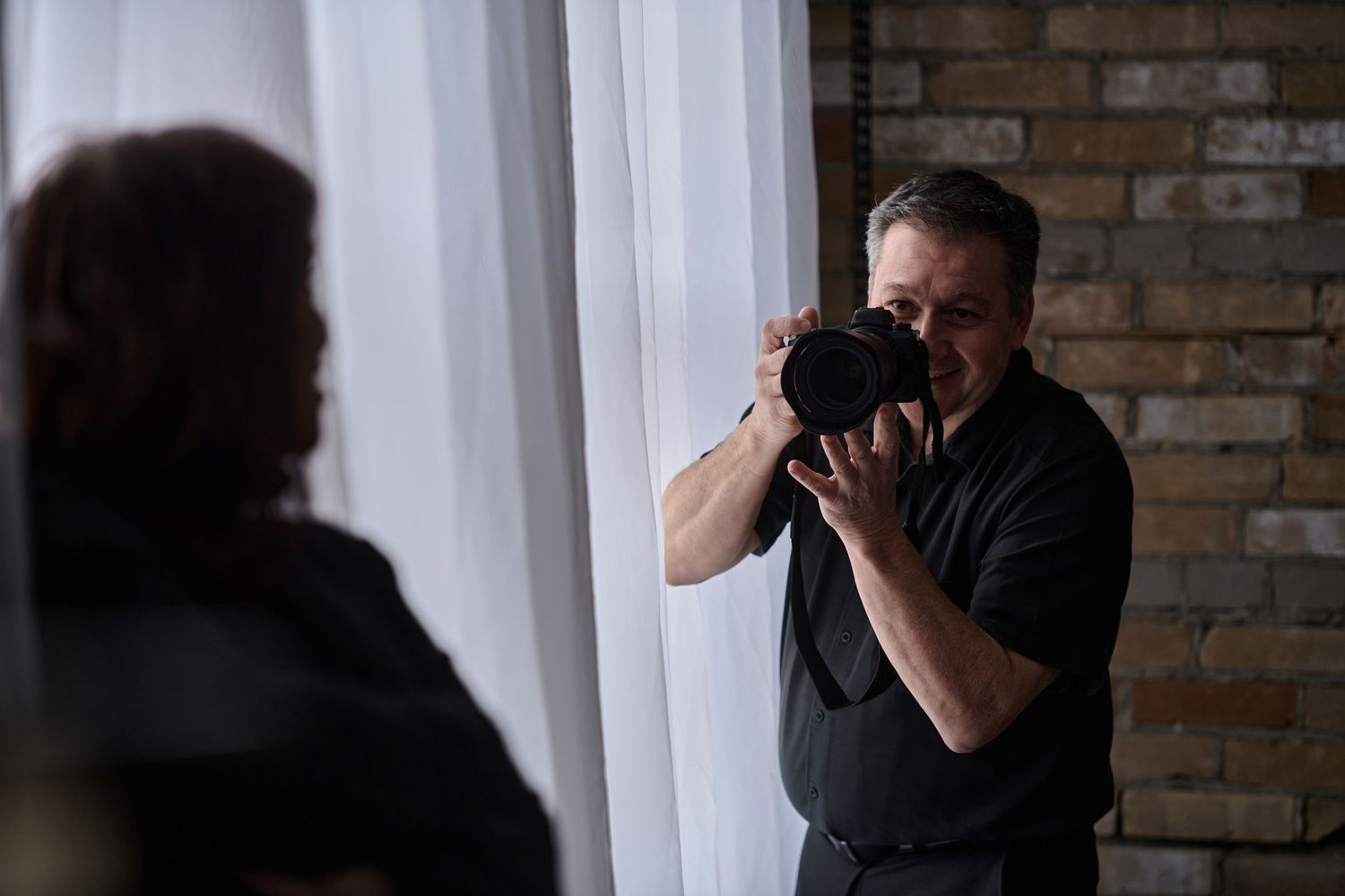 A photographer in dark clothing takes photos near a brick wall and white curtain.