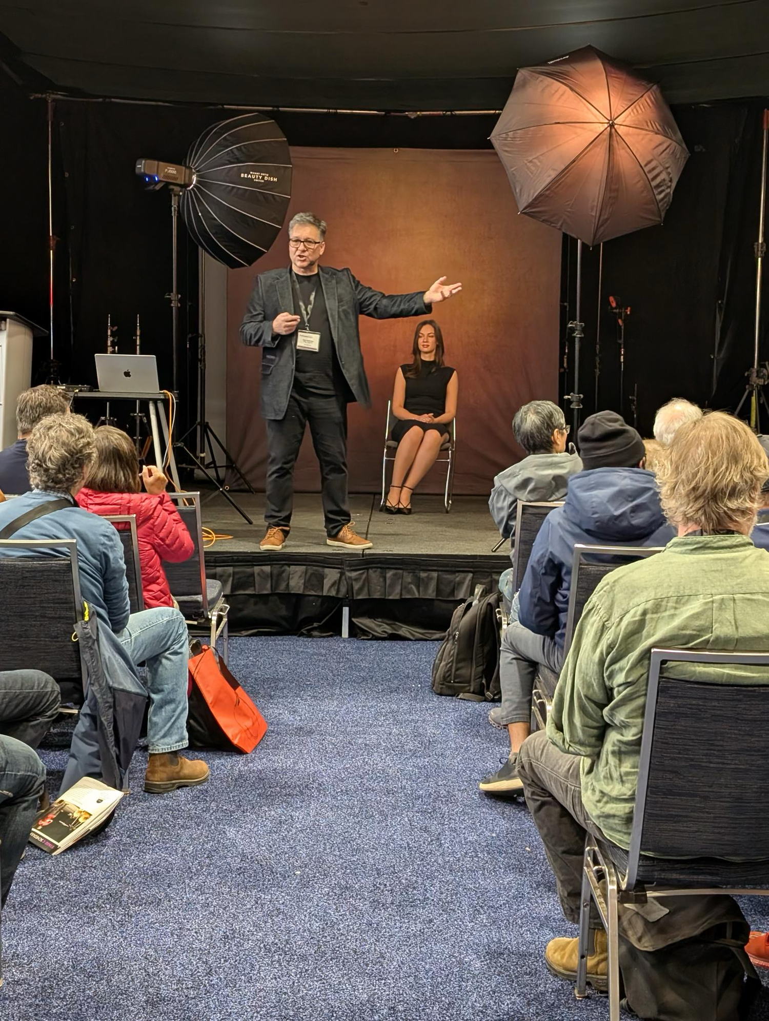 Photography studio setup with lighting equipment and audience members seated on blue carpet.