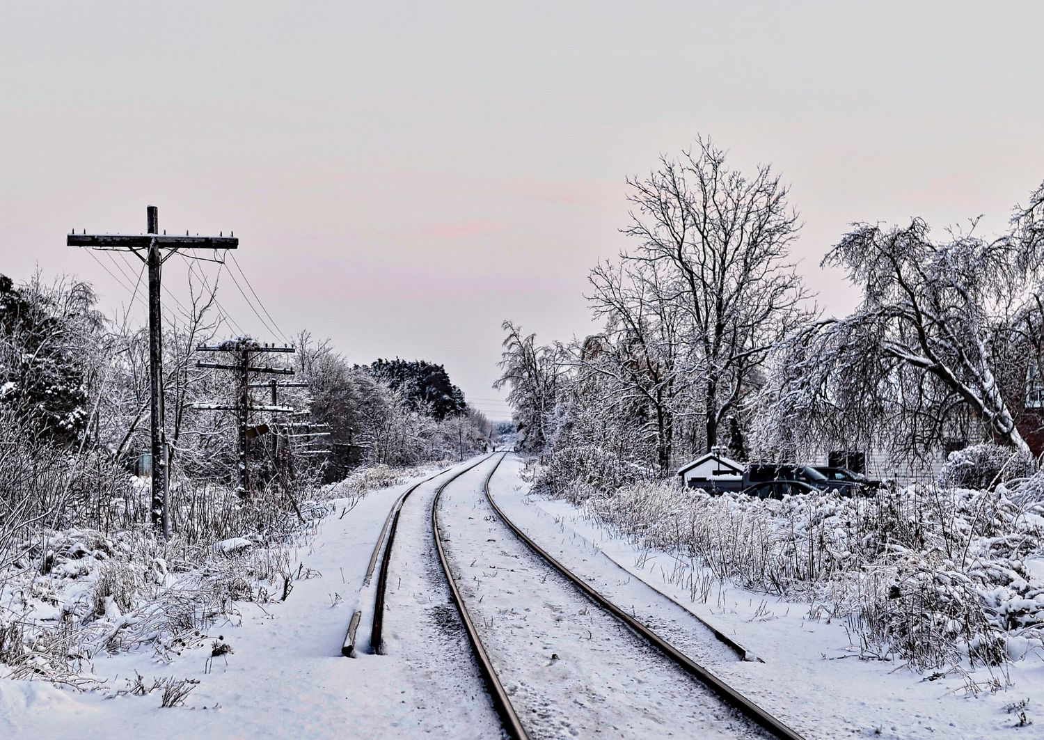 Snow covered railroad tracks converge into the distance through a winter landscape with bare trees and utility poles.