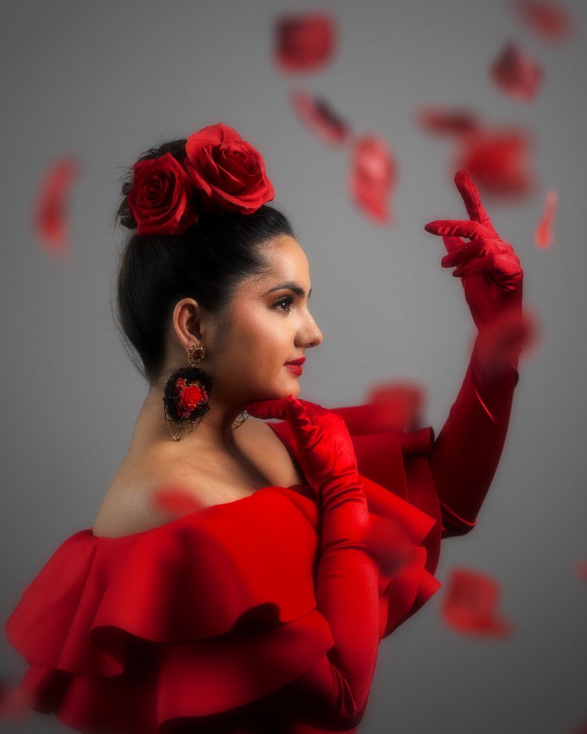 A dancer in a flowing red dress with matching gloves poses elegantly against a gray background with floating rose petals.