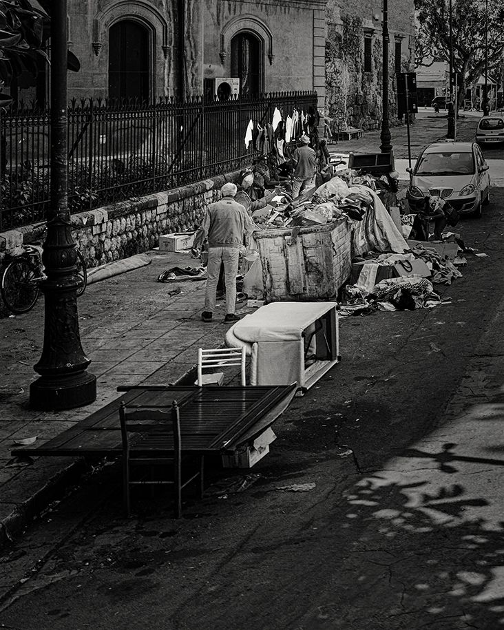 Black and white photo of people resting on benches along an urban sidewalk with trees casting shadows.