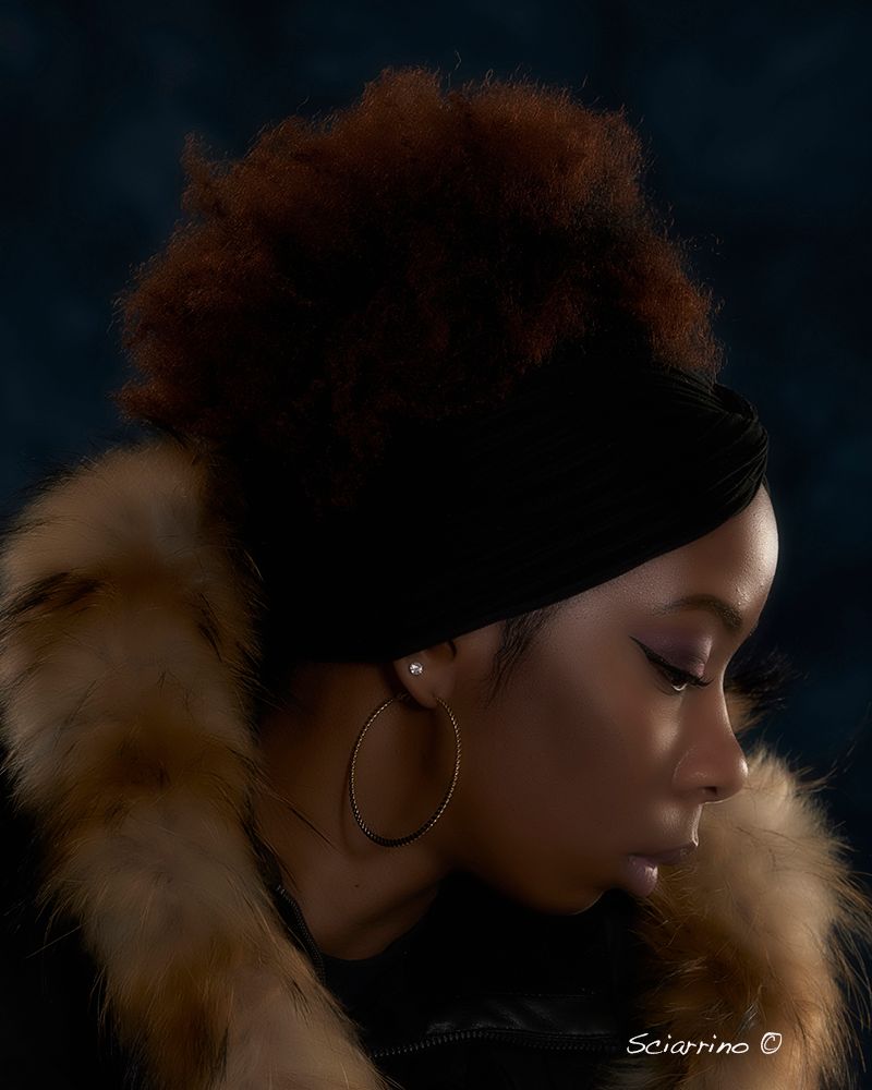 Artistic profile portrait with dramatic dark lighting and warm tones highlighting curly hair and fur collar details.