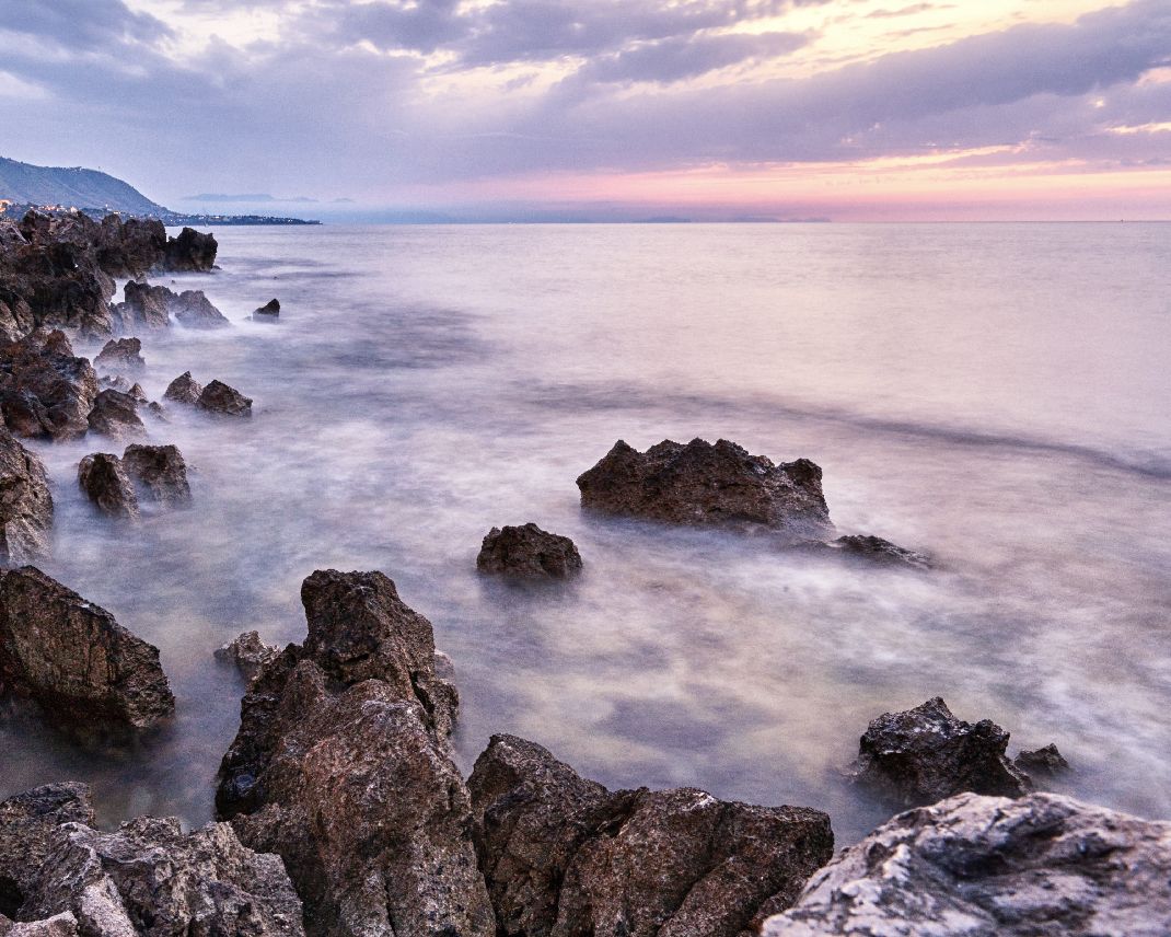 Rocky coastline with waves crashing against jagged rocks during sunset creating a misty seascape.