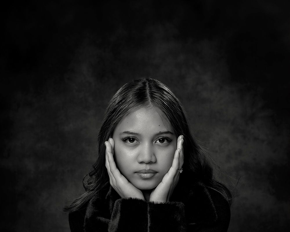 Black and white portrait of a young woman holding her face with both hands against a dark background.