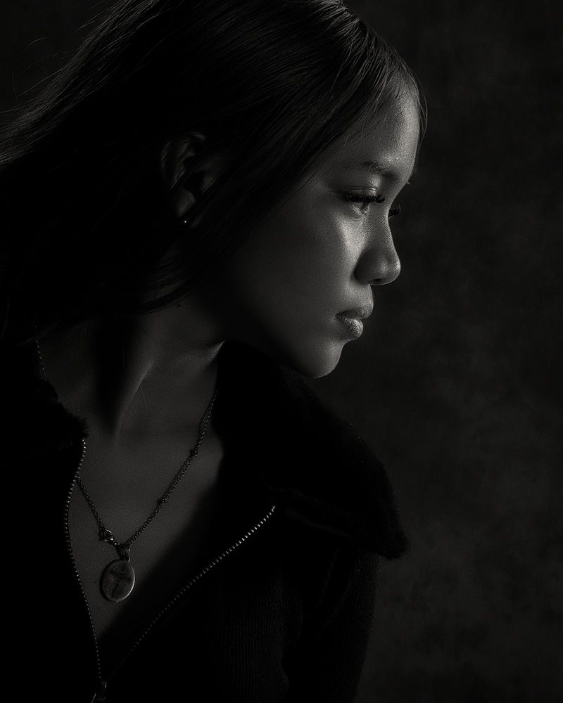 Dramatic black and white profile portrait with moody lighting highlighting delicate features and a pendant necklace.