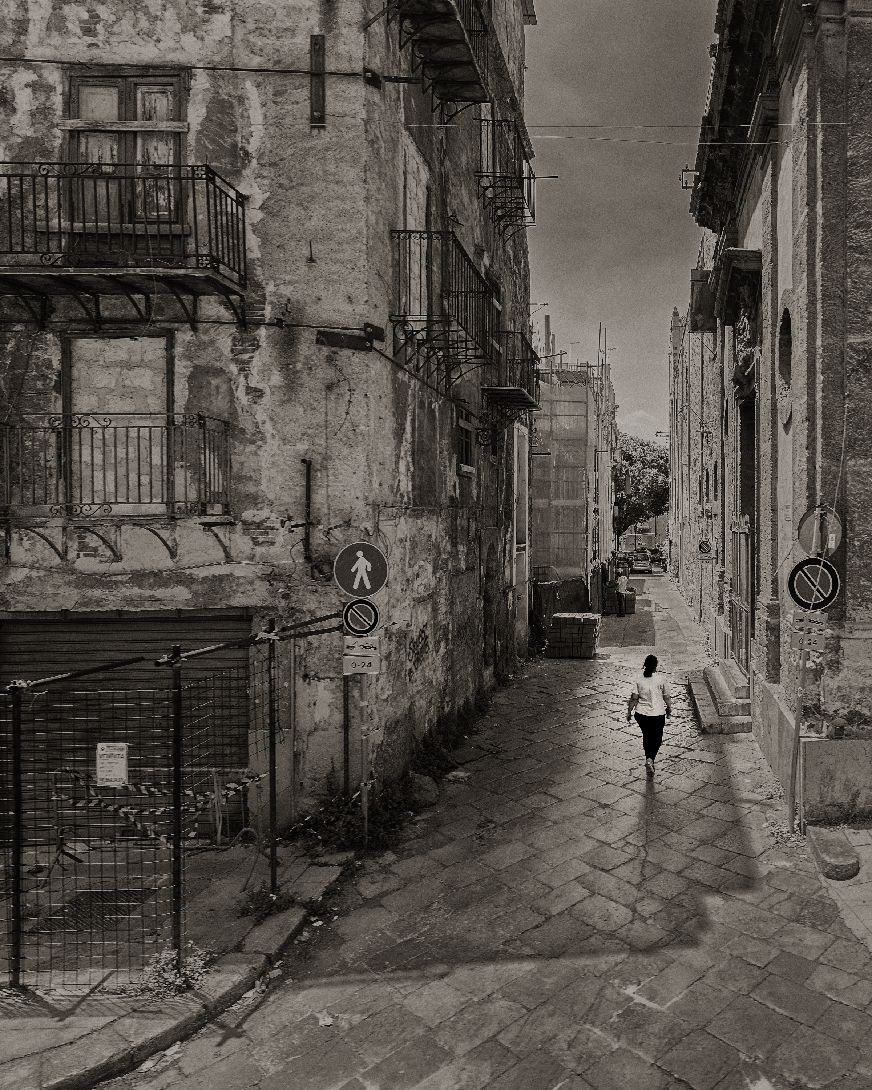 A lone figure walks down a narrow cobblestone street between old weathered buildings in a black and white photograph.
