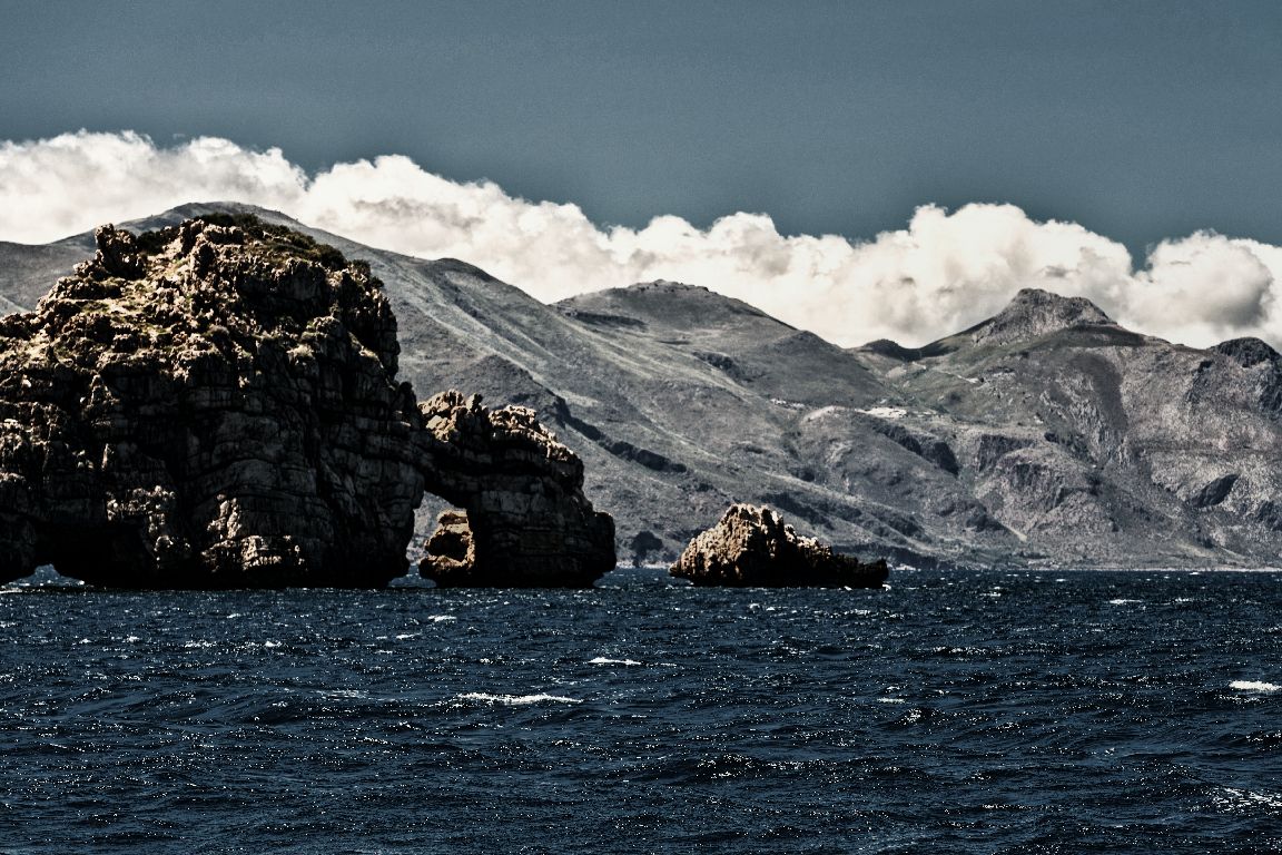 Scenic mountain landscape with rocky cliffs rising above dark blue ocean waters under a cloudy sky.
Horvath-Photography-Sam-Sciarrino