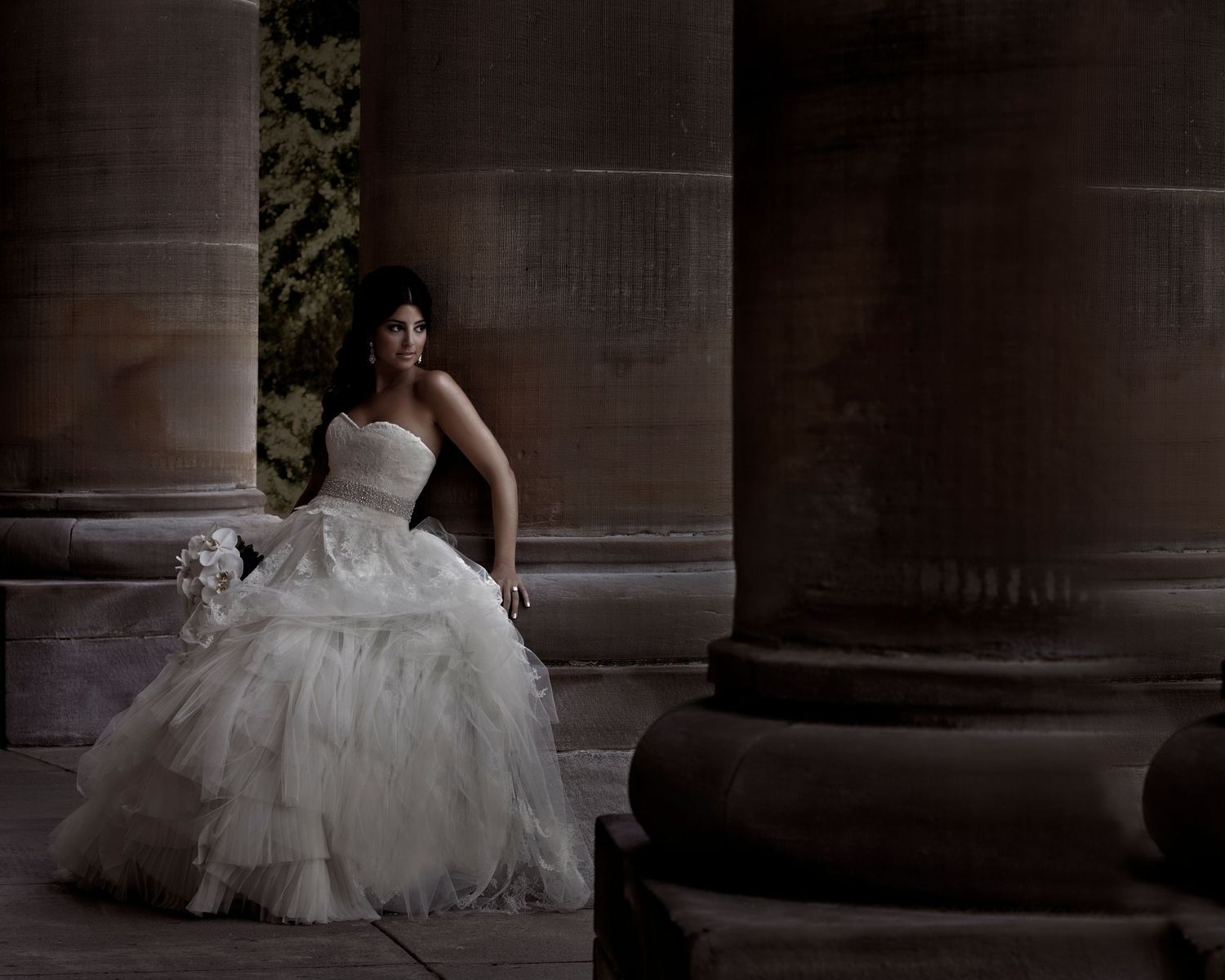 A bride in a layered white tulle wedding gown poses elegantly against stone columns in dramatic lighting.