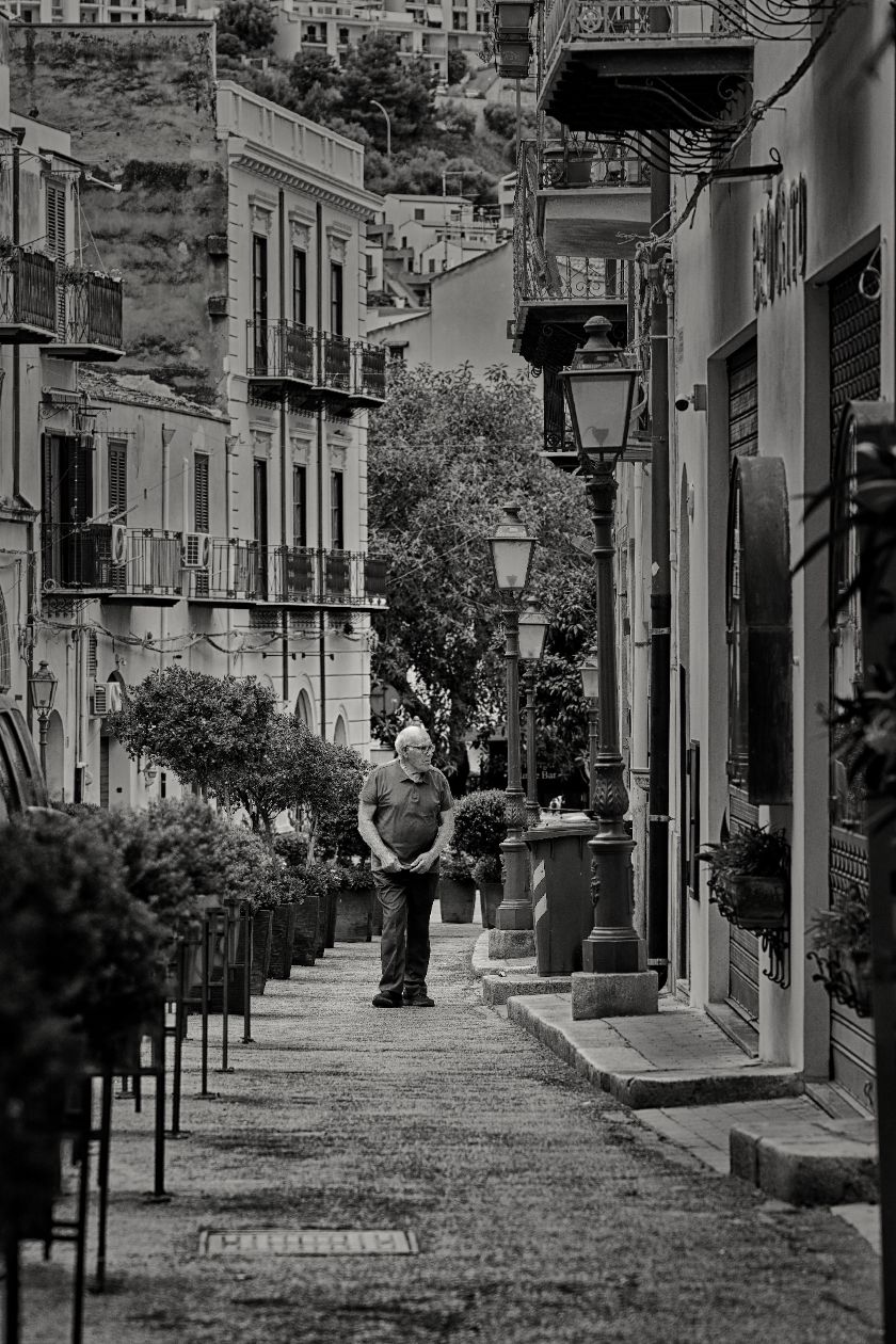 A black and white photograph of a narrow cobblestone street lined with historic buildings and lamp posts.