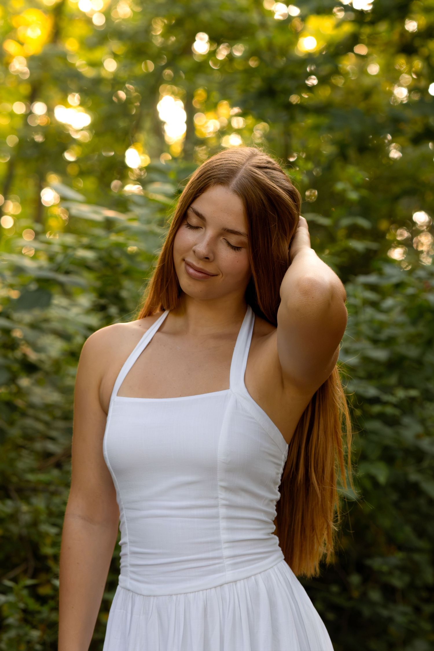 Young woman with long auburn hair in white halter dress poses outdoors among lush green trees in golden sunlight.