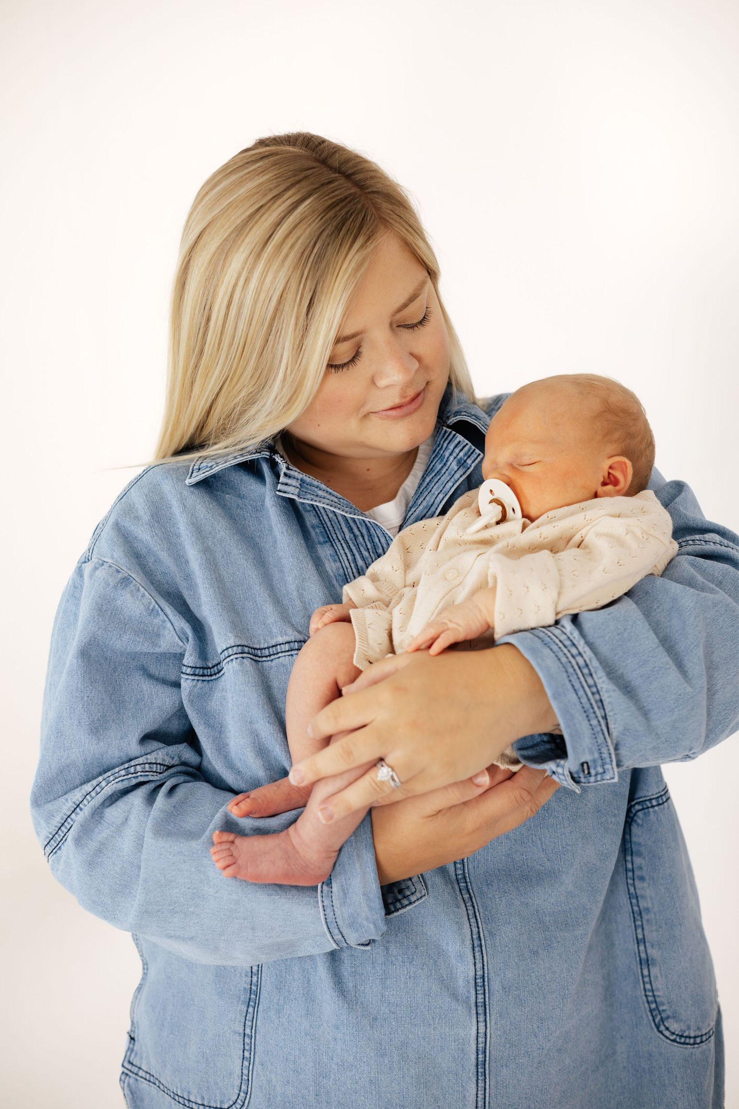 Blonde woman in denim jacket tenderly holding a sleeping newborn baby against a white background.