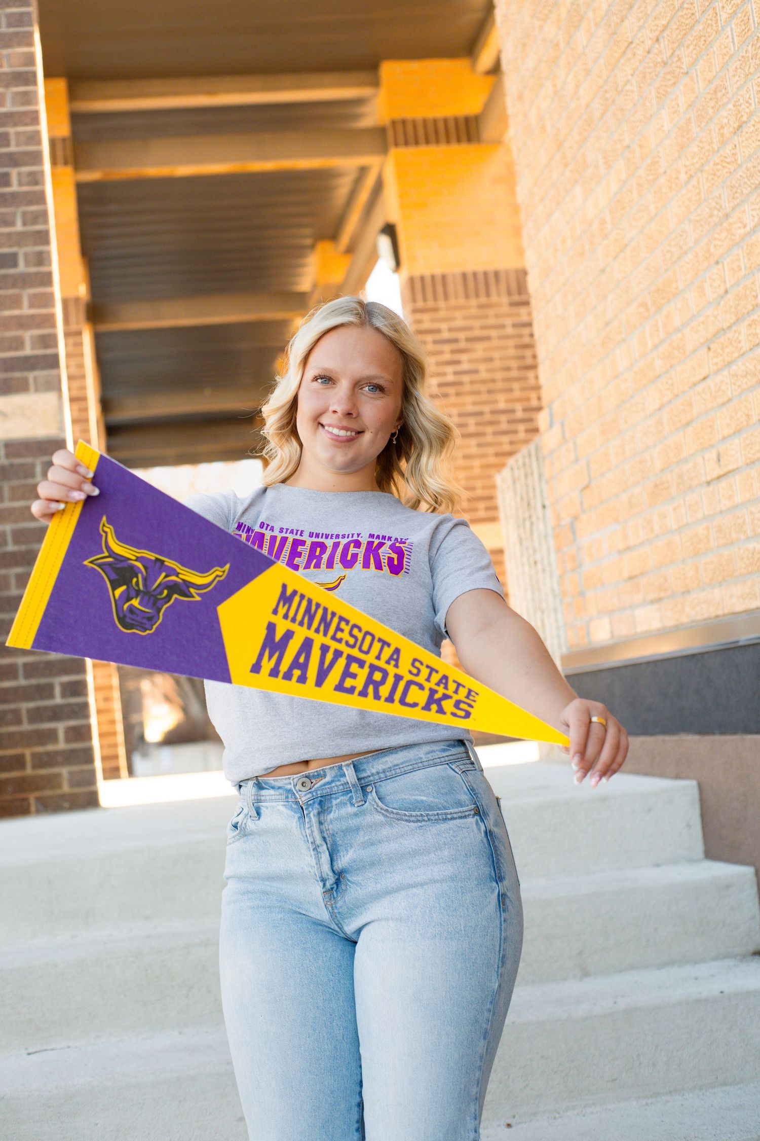 Smiling blonde woman holding Minnesota State Mavericks pennants on campus steps.