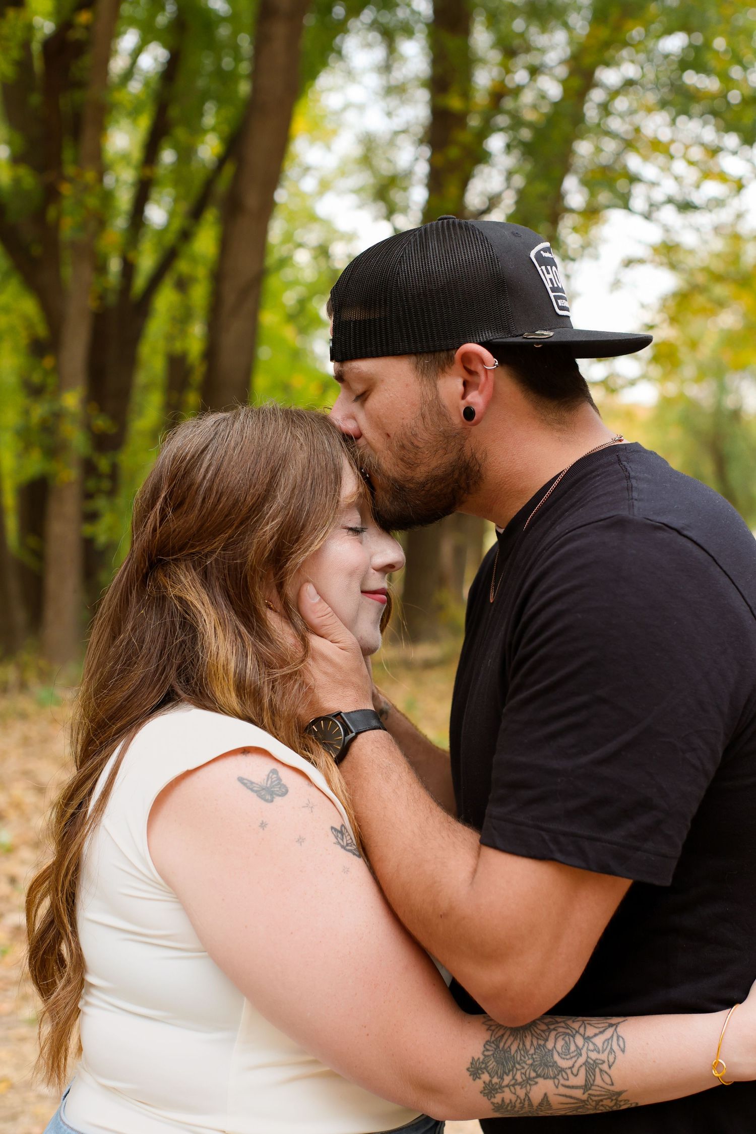 A couple shares an intimate moment outdoors in a wooded setting with autumn trees in the background.