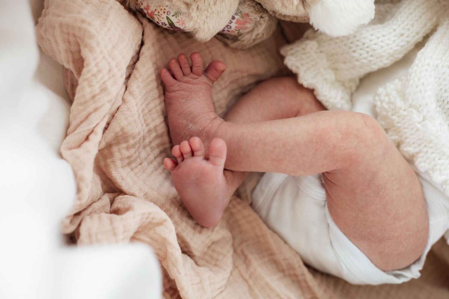 Close-up of newborn baby feet resting on a soft beige muslin blanket with a white knit toy nearby.