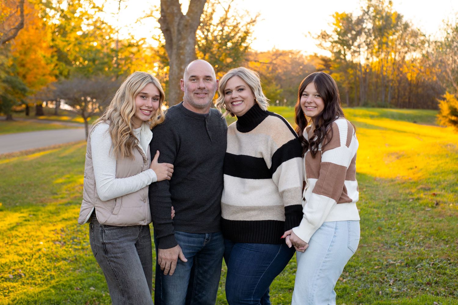Family of four posing together outdoors in a sunlit park, wearing cozy sweaters and jeans, with golden autumn trees in the background.