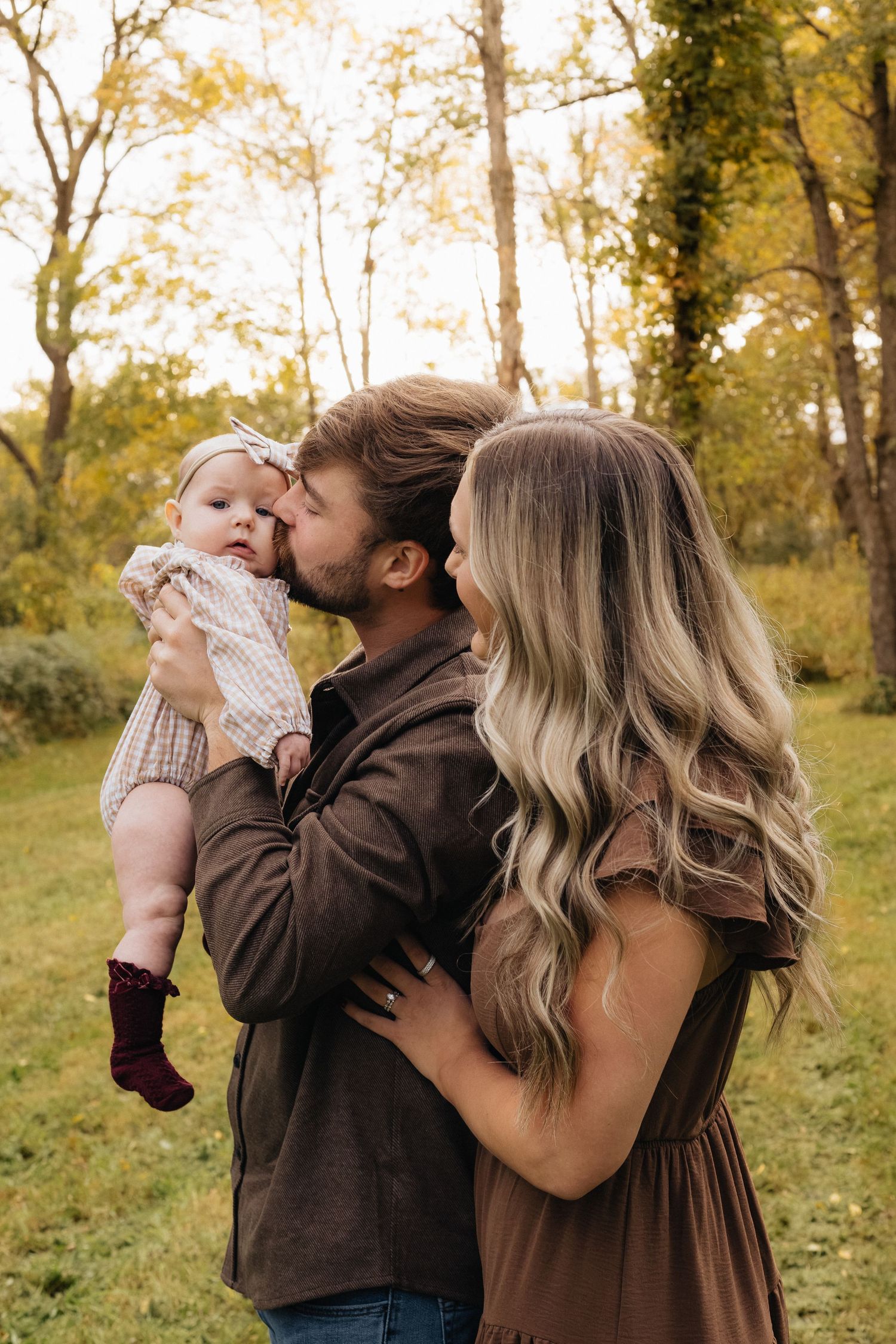 A man and woman embrace while the man kisses a baby in a headband and burgundy boots, standing in a sunny autumn park with golden trees.