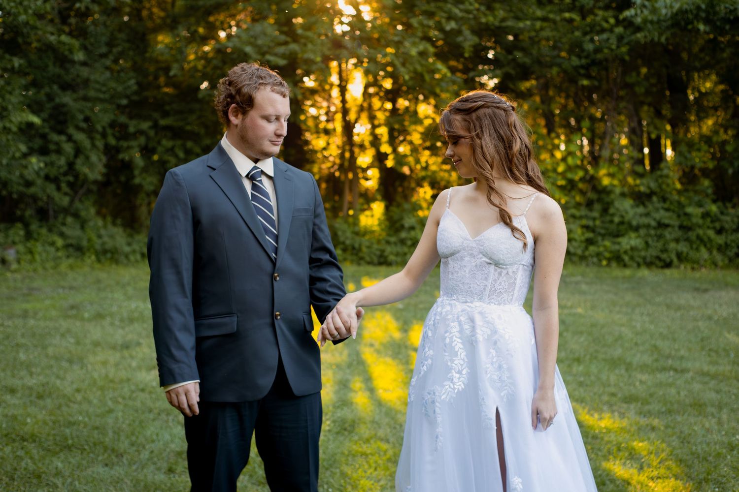 Bride in white lace gown and groom in gray suit hold hands outdoors at golden hour wedding ceremony.