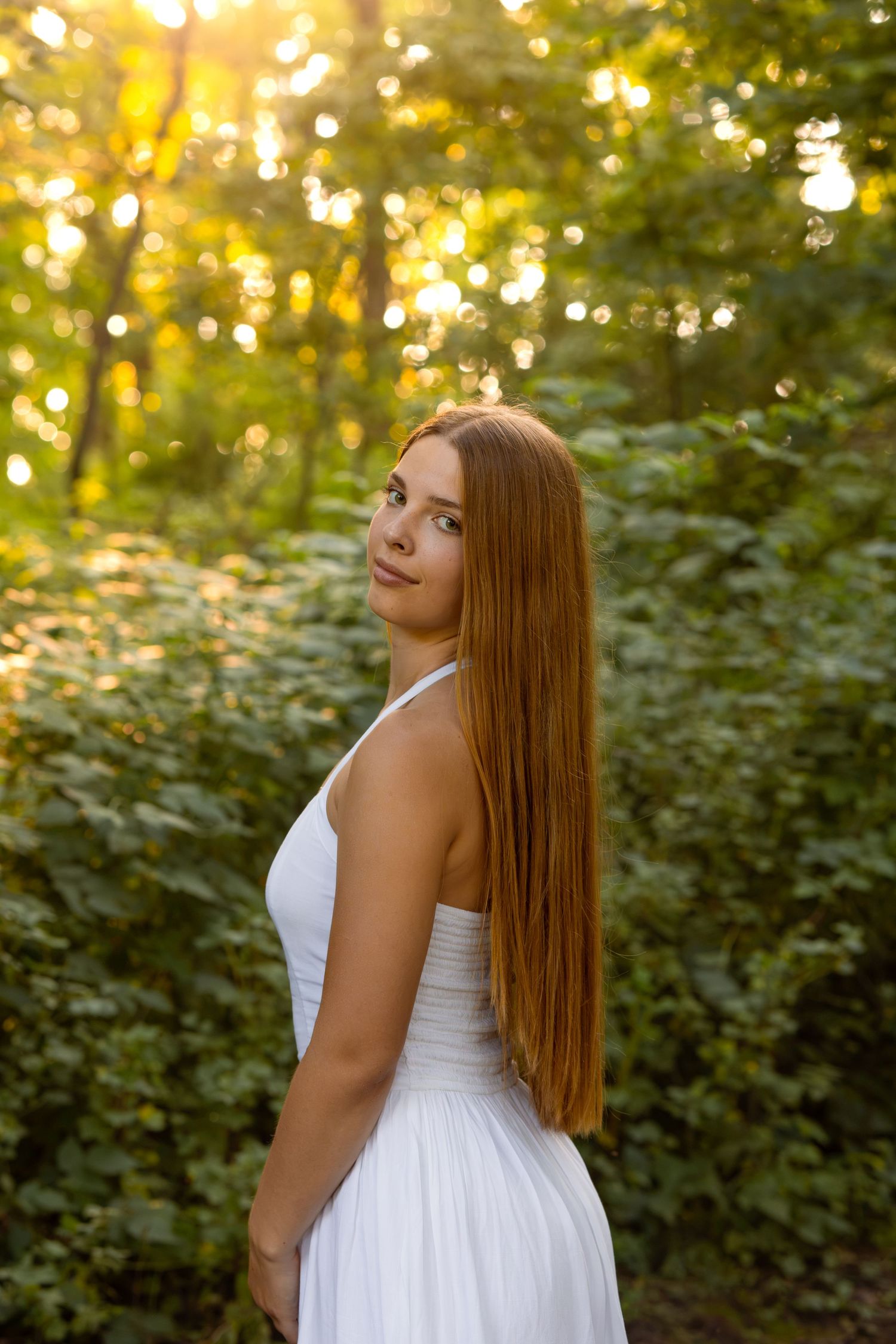 Young woman with long red hair in white dress posing in sunlit forest with green foliage background.
