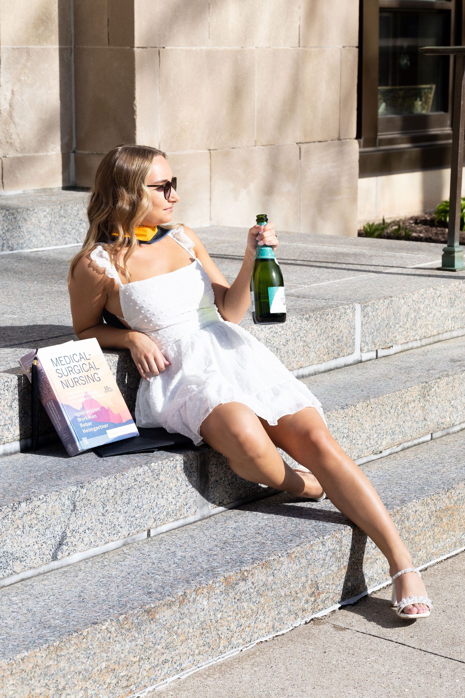 Young woman in white dress and sunglasses sits on steps celebrating graduation with champagne and nursing textbook.