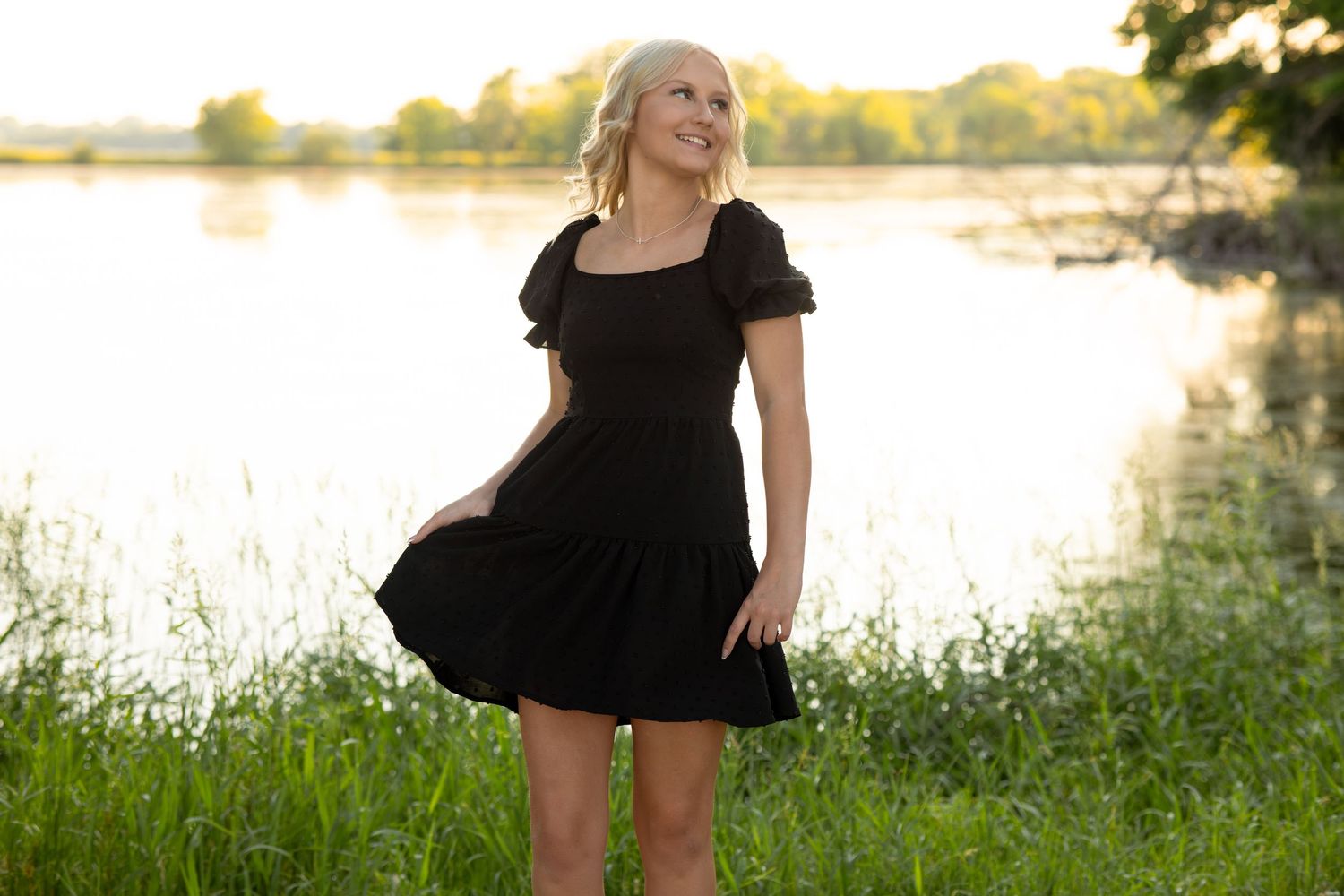 Blonde woman in black mini dress standing by a lake in green grass, smiling in golden hour sunlight.