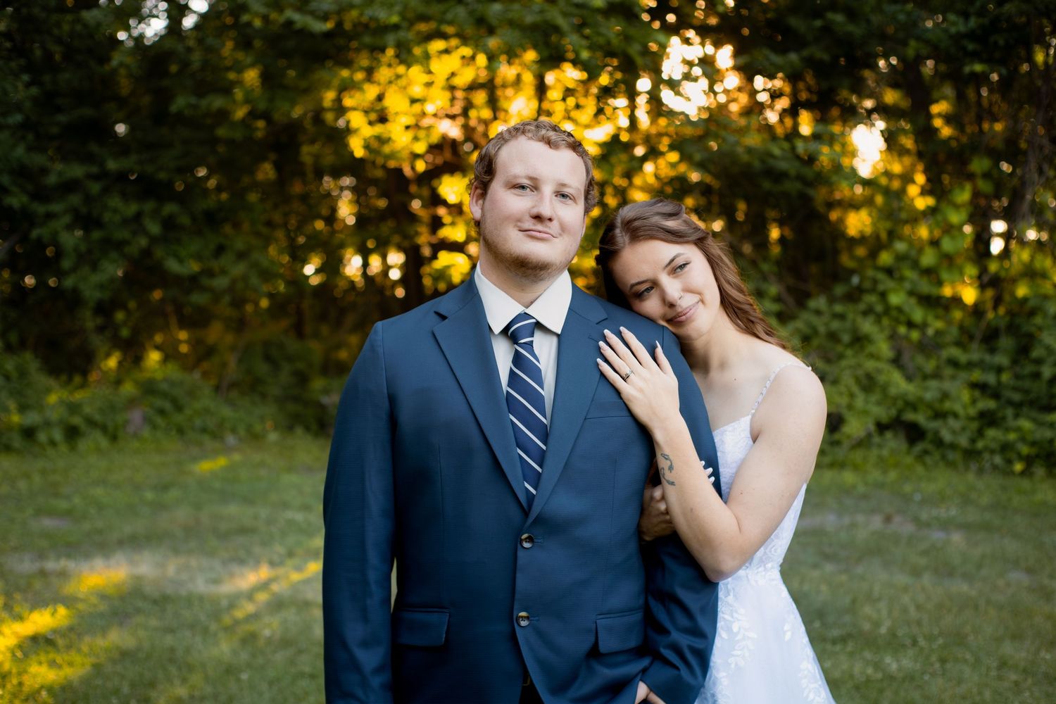 Couple posing outdoors in golden hour light, man in navy suit and woman in white dress leaning on his shoulder.