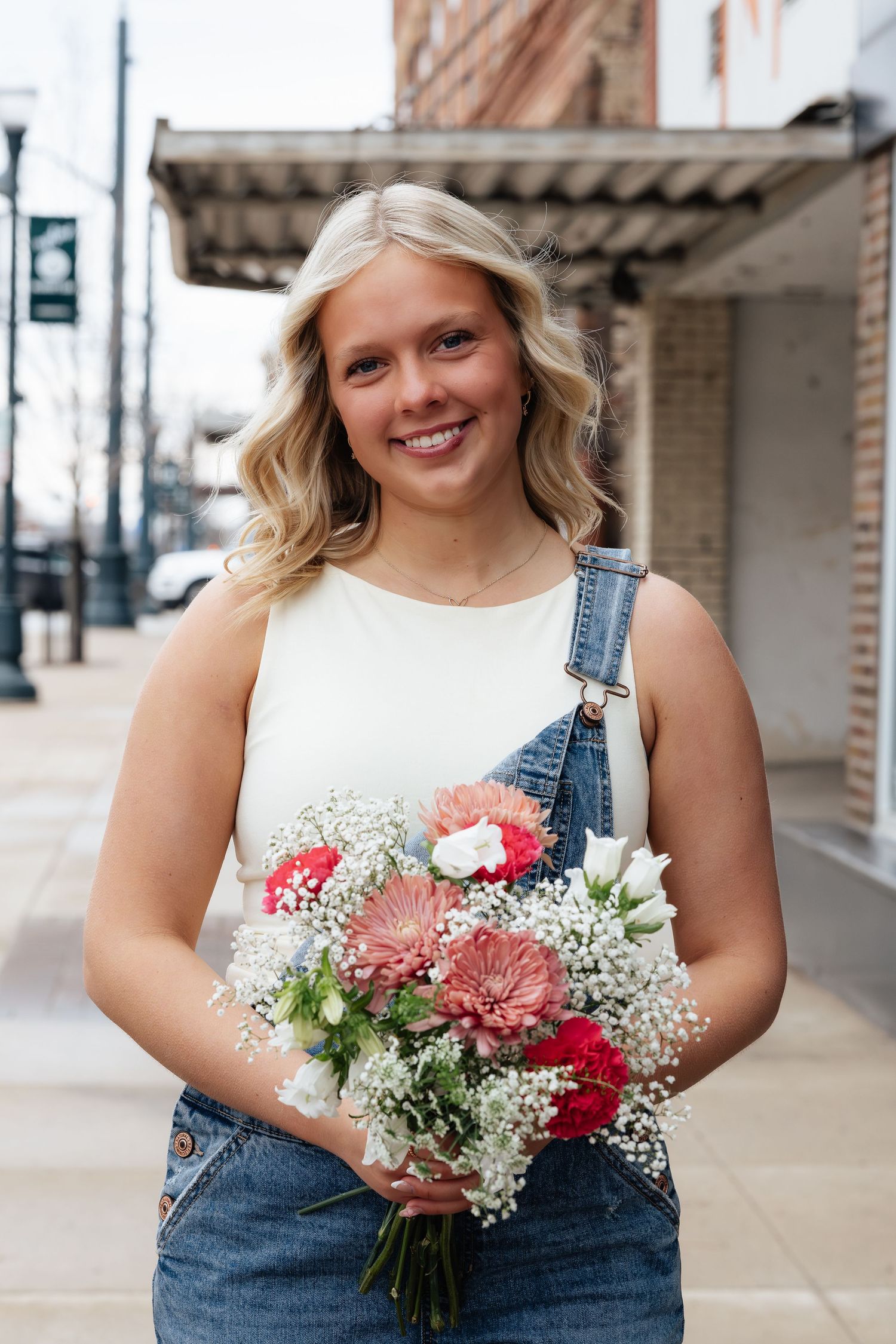Smiling blonde woman in denim overalls holding a bouquet of pink and white flowers on a city sidewalk.