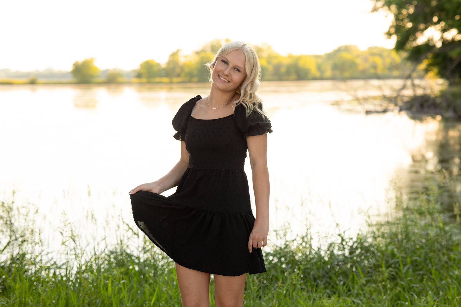 Smiling blonde woman in black dress standing by a sunlit lake surrounded by lush green grass.