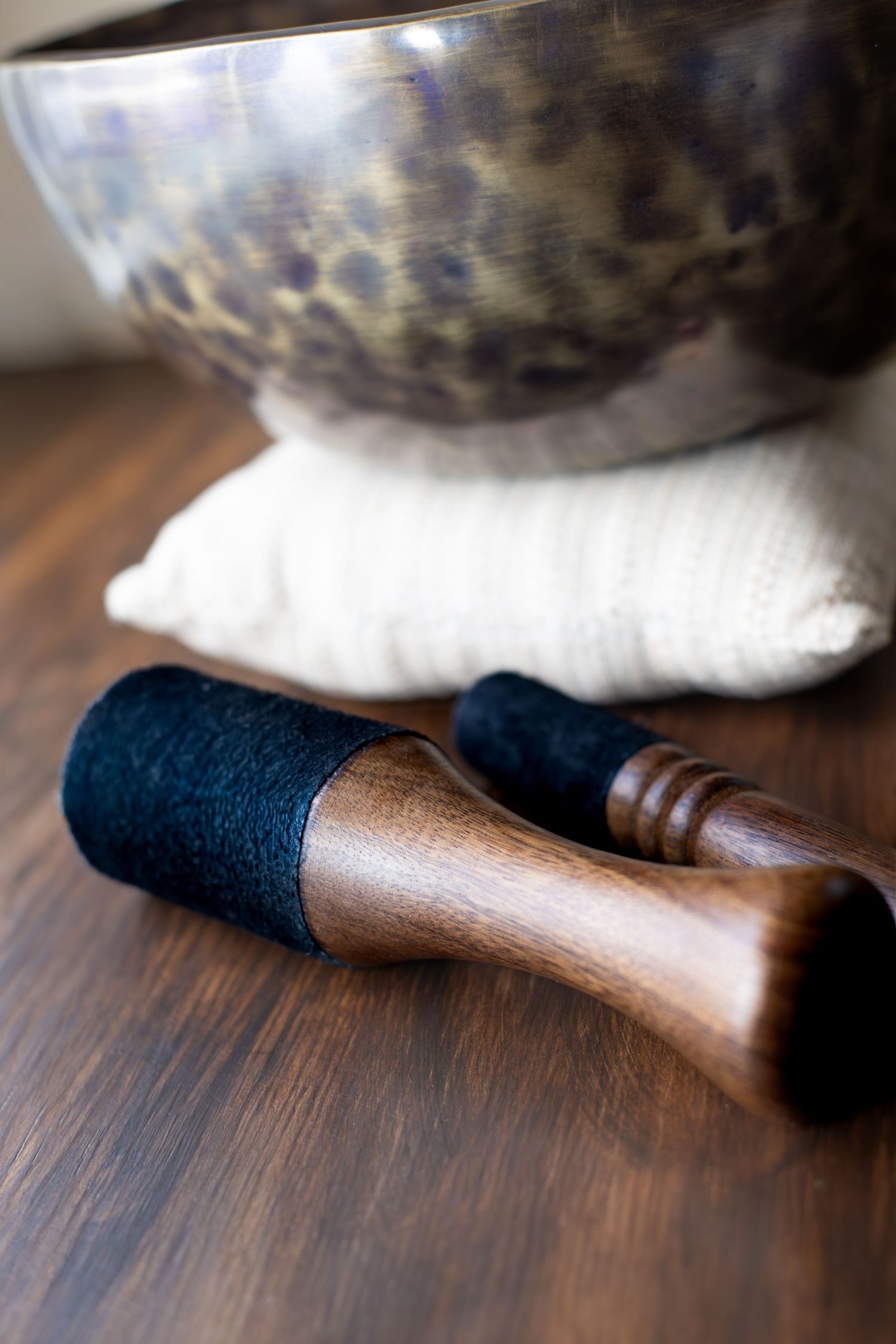 Tibetan singing bowl resting on white cushion with wooden mallet on dark wood surface.
