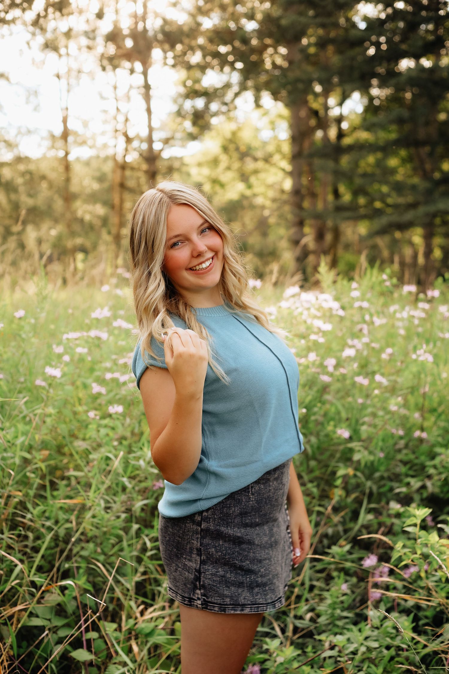 A woman in blue top and denim skirt poses in a wildflower meadow at sunset.