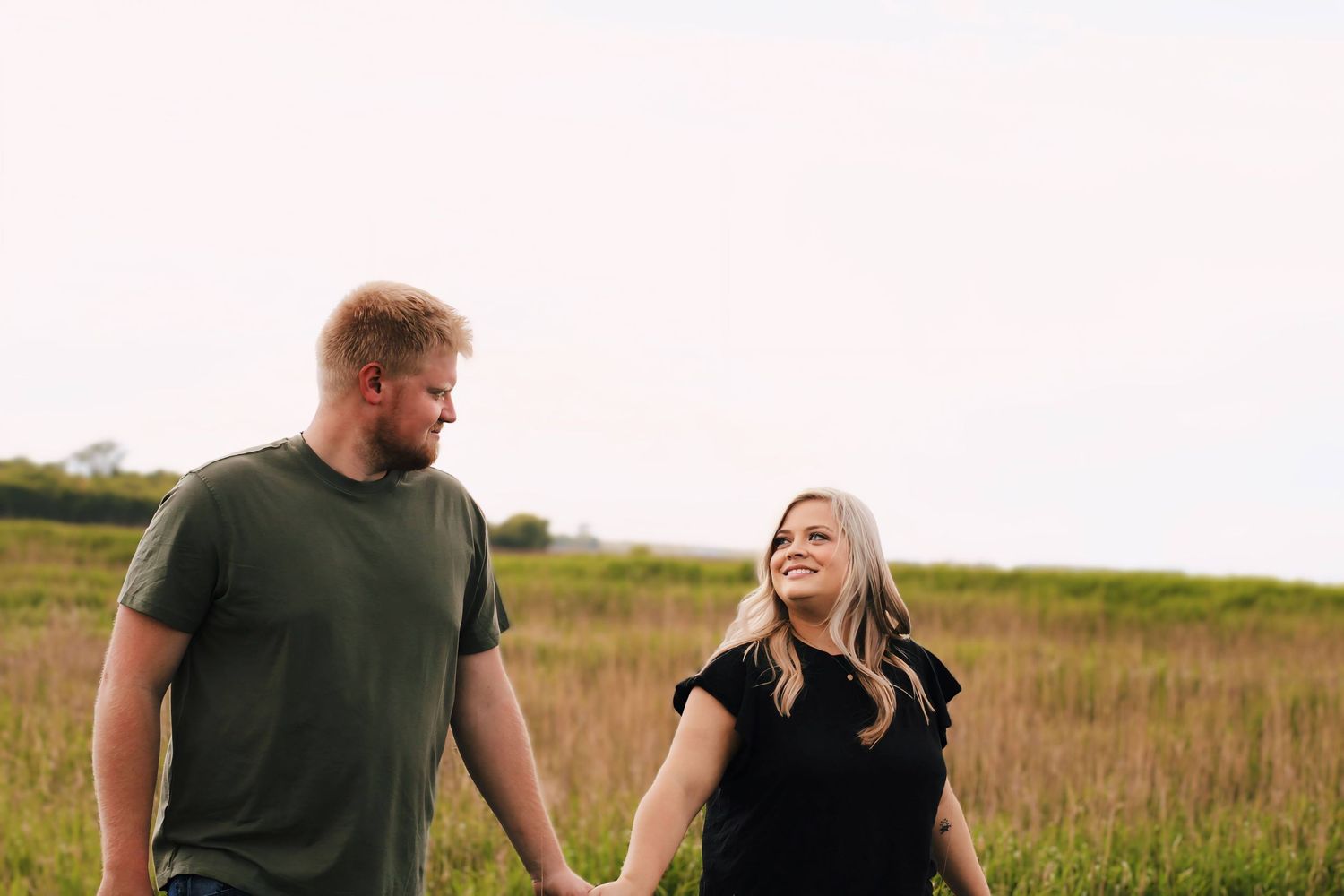 Couple walking and laughing together through a tall grass field at sunset.
