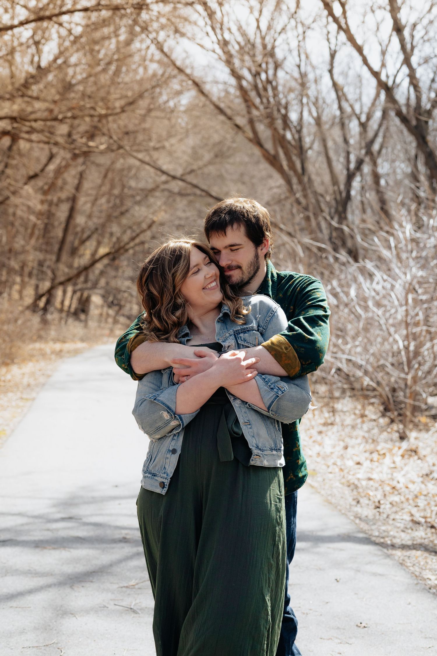 Winter portrait of couple embracing on snowy path surrounded by bare trees.