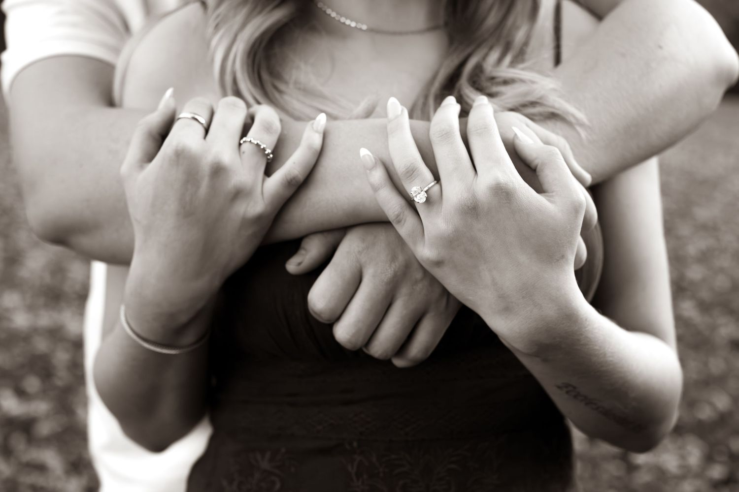 Black and white photo of a person embracing a woman from behind, her hands with rings resting on their arms in a romantic engagement pose.