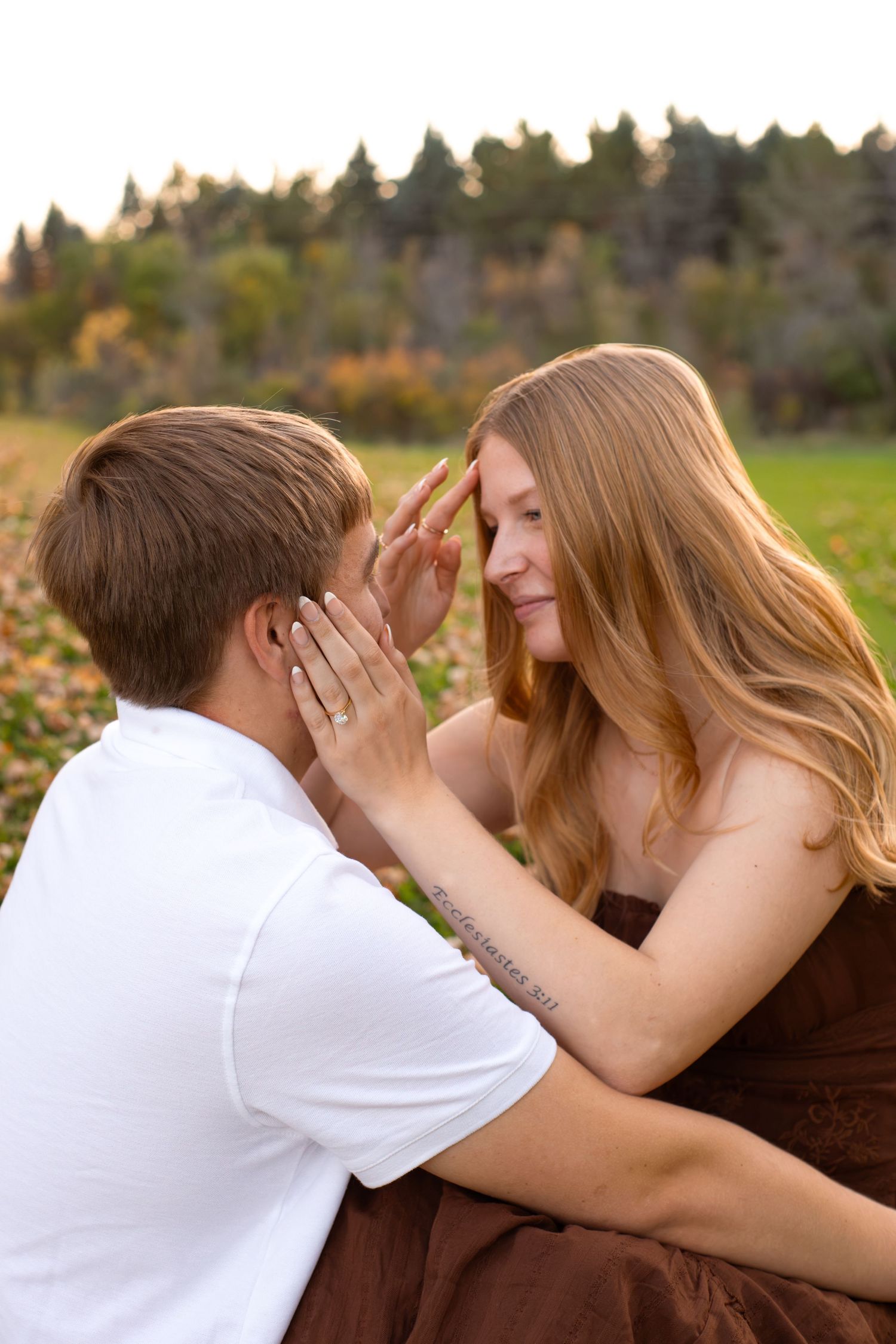 Couple sharing an intimate moment outdoors as a woman with long auburn hair touches foreheads with a man in a white shirt at autumn sunset.