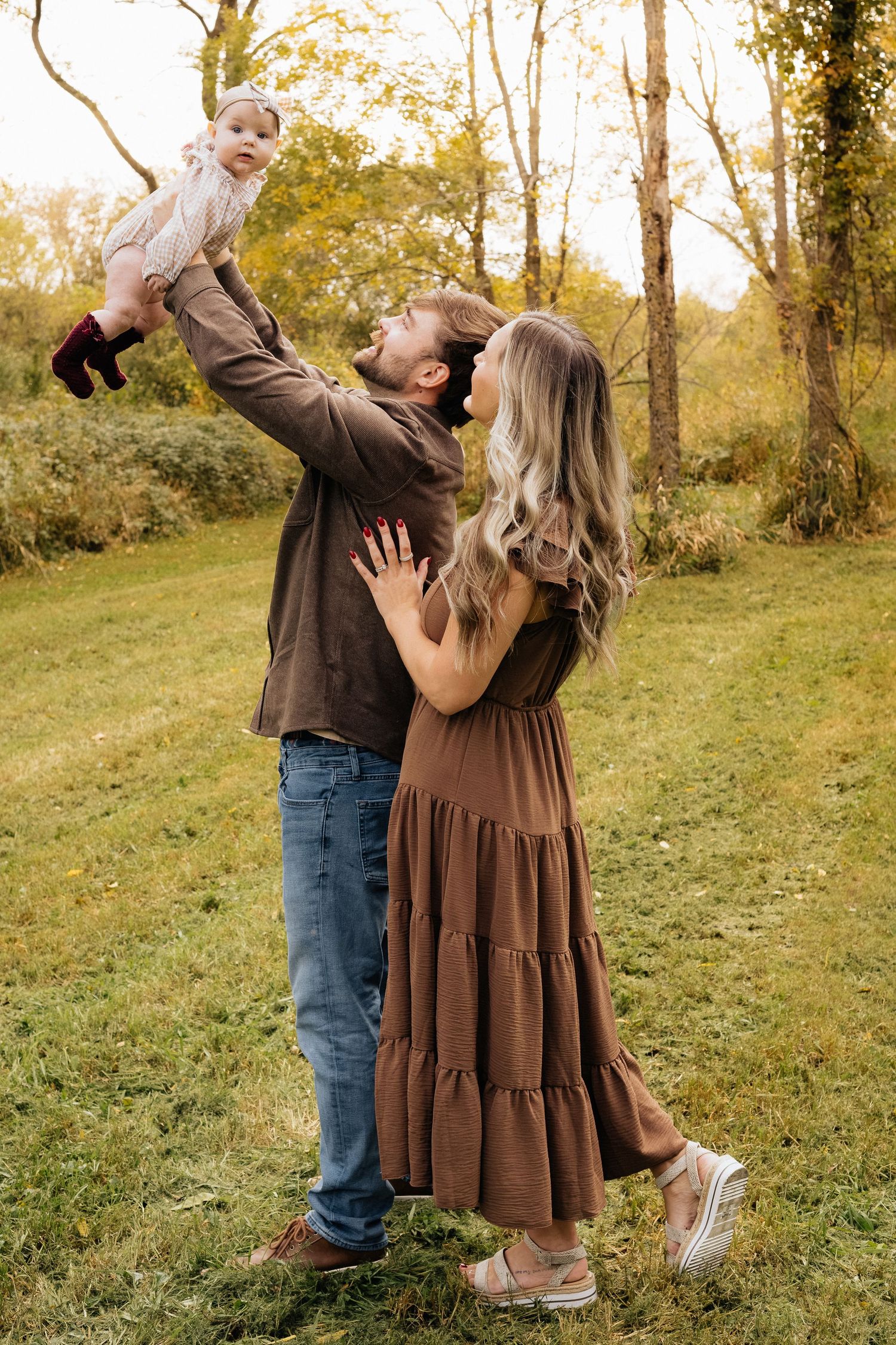 A smiling man lifts a baby into the air beside a woman in a brown tiered dress on a grassy field with autumn trees.