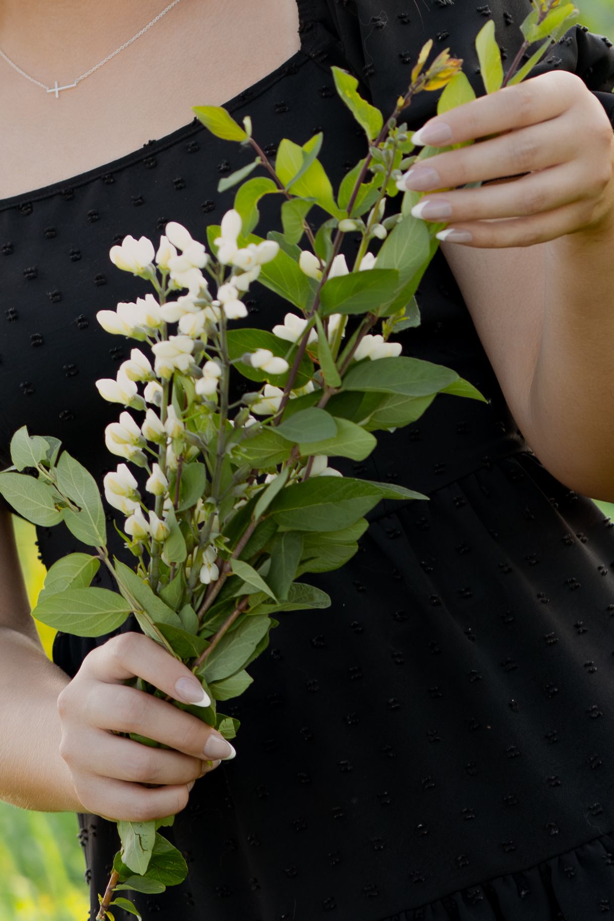 Woman in black dress holding white wildflowers and green leaves bouquet outdoors.