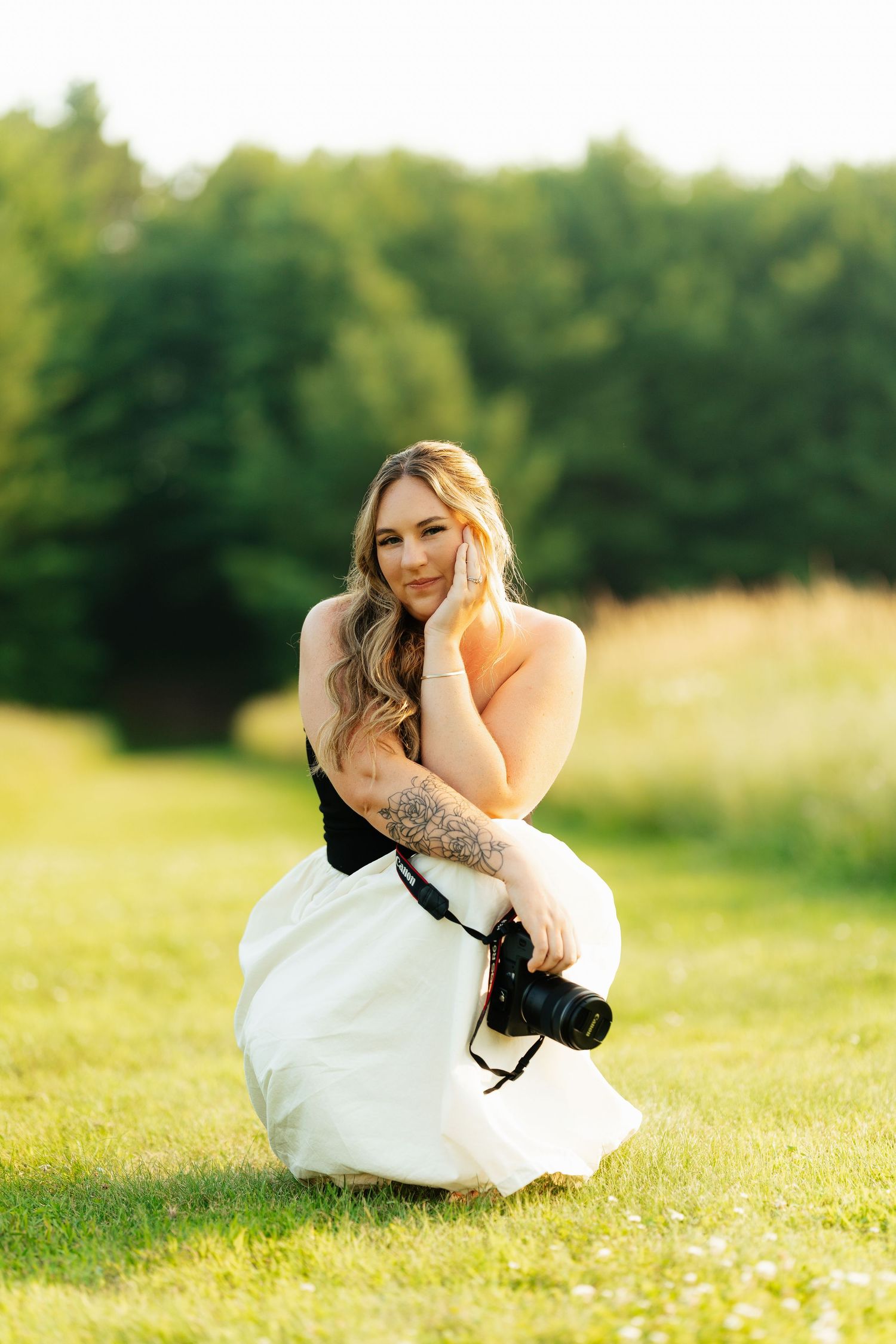 Photographer in white dress kneeling with camera in sunny meadow.