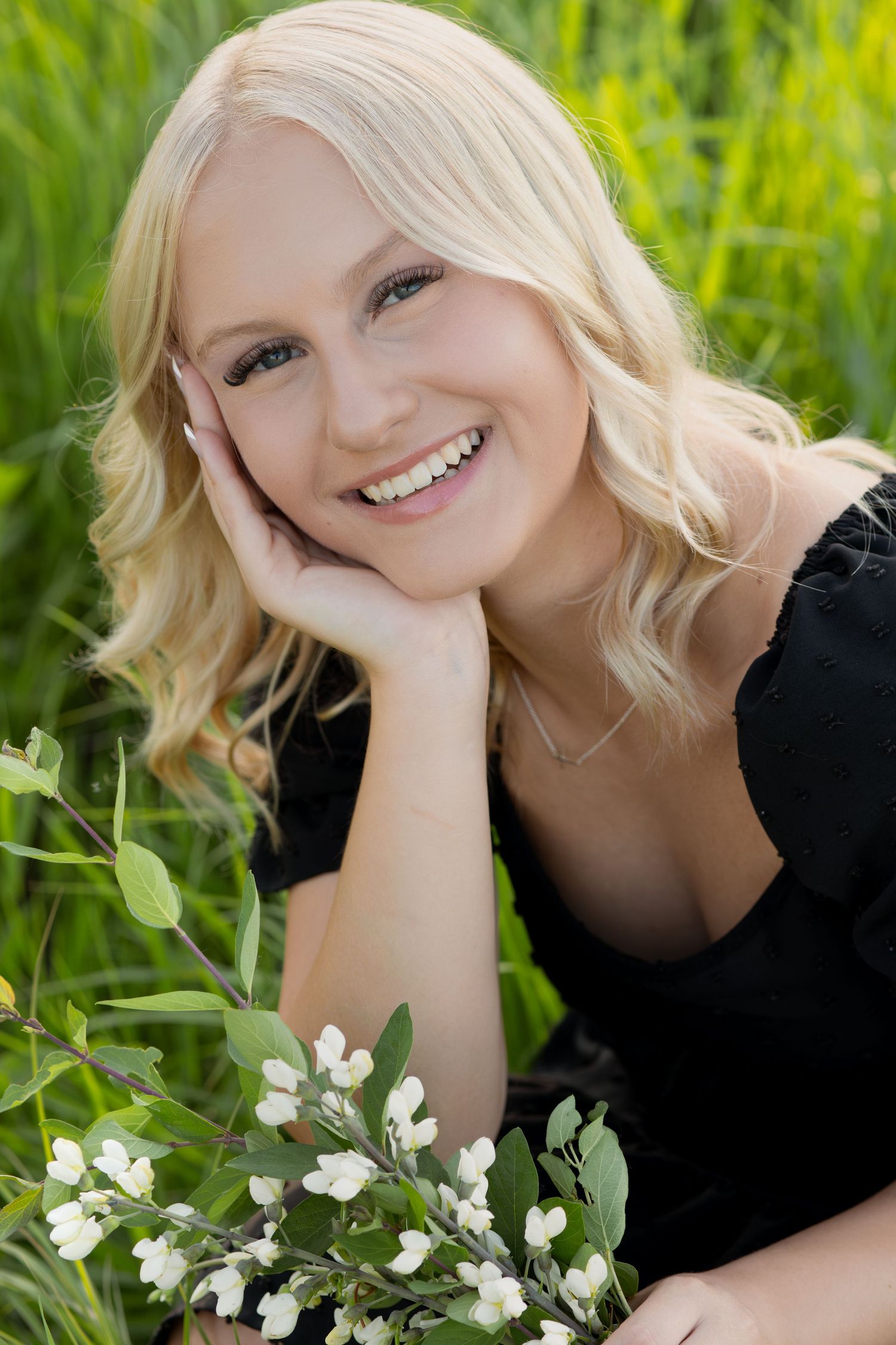 Smiling blonde woman in black dress resting in green grass field holding white wildflowers on a sunny day.
