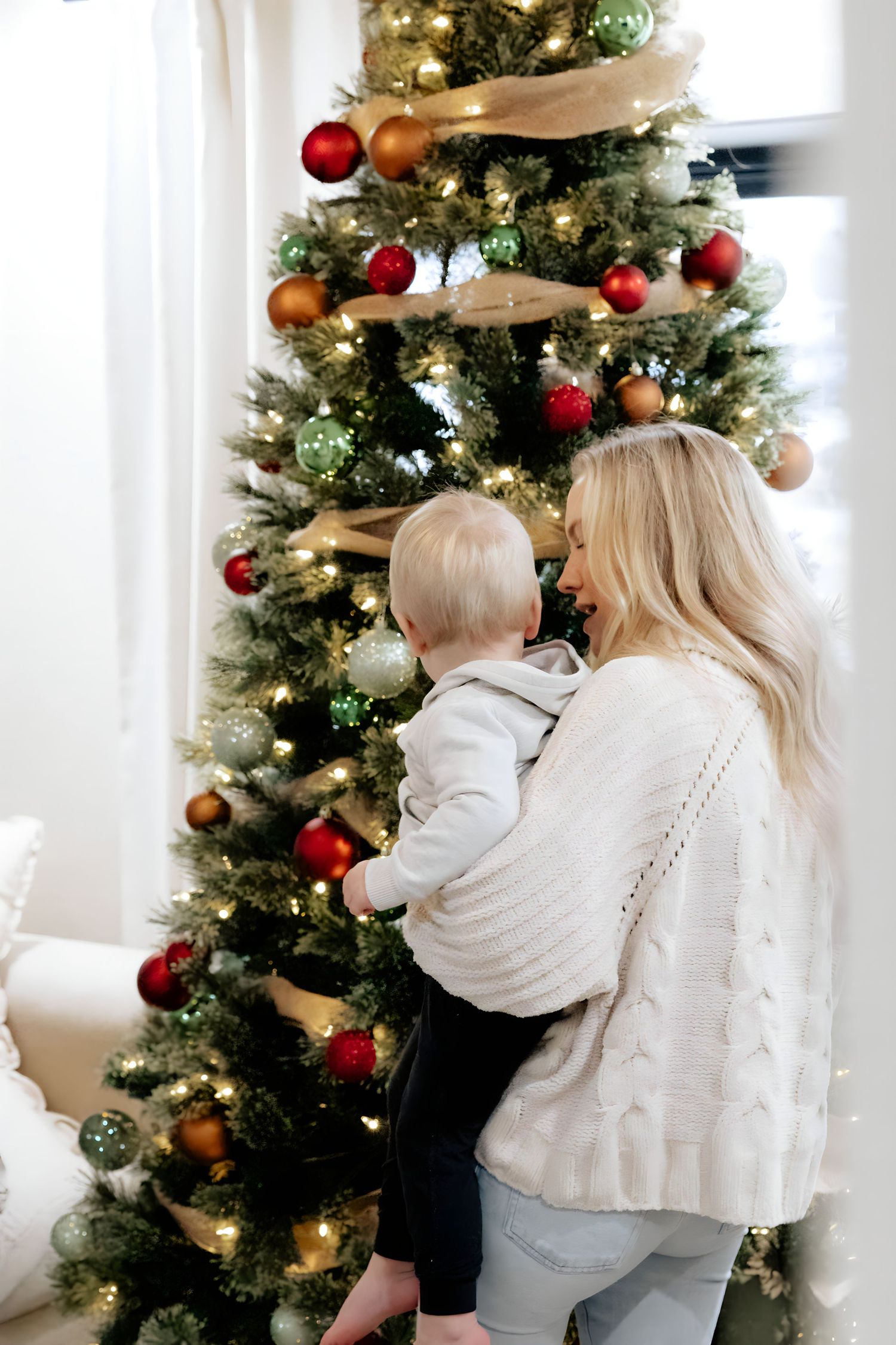 Person holding child while decorating Christmas tree with red and gold ornaments.