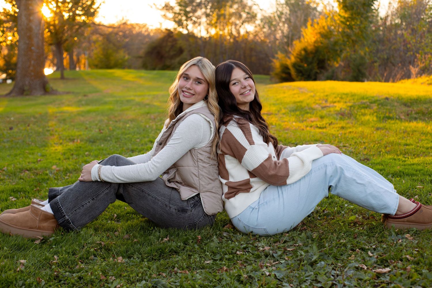 Two young women sit back to back on a sunny grassy lawn, smiling in casual fall outfits with golden-hour light and trees behind them.