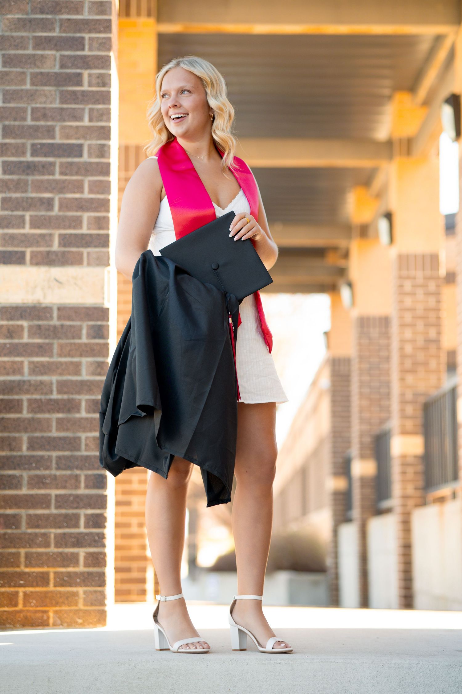 Smiling blonde graduate holding cap and gown in white dress with red sash, posing outdoors near brick building.