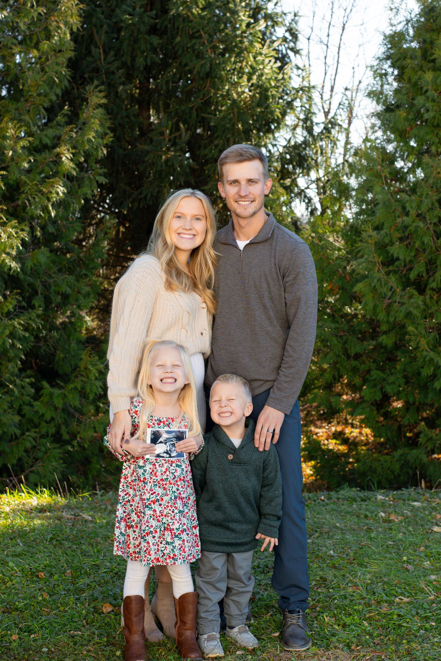 A happy family of four poses outdoors among evergreen trees, with two young children smiling in front of their parents.