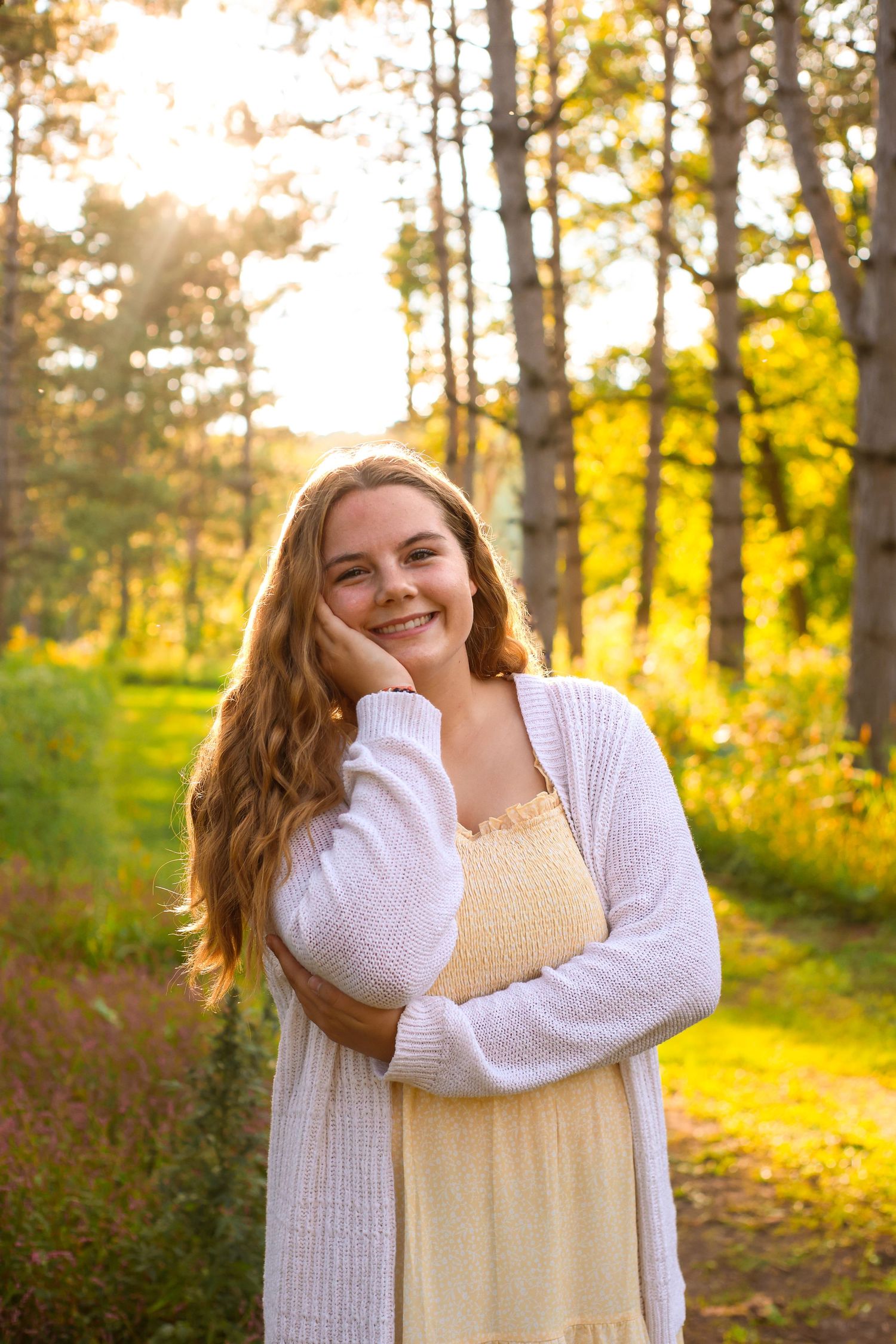 A person in a white sweater and yellow dress poses outdoors during golden hour in a wooded setting.