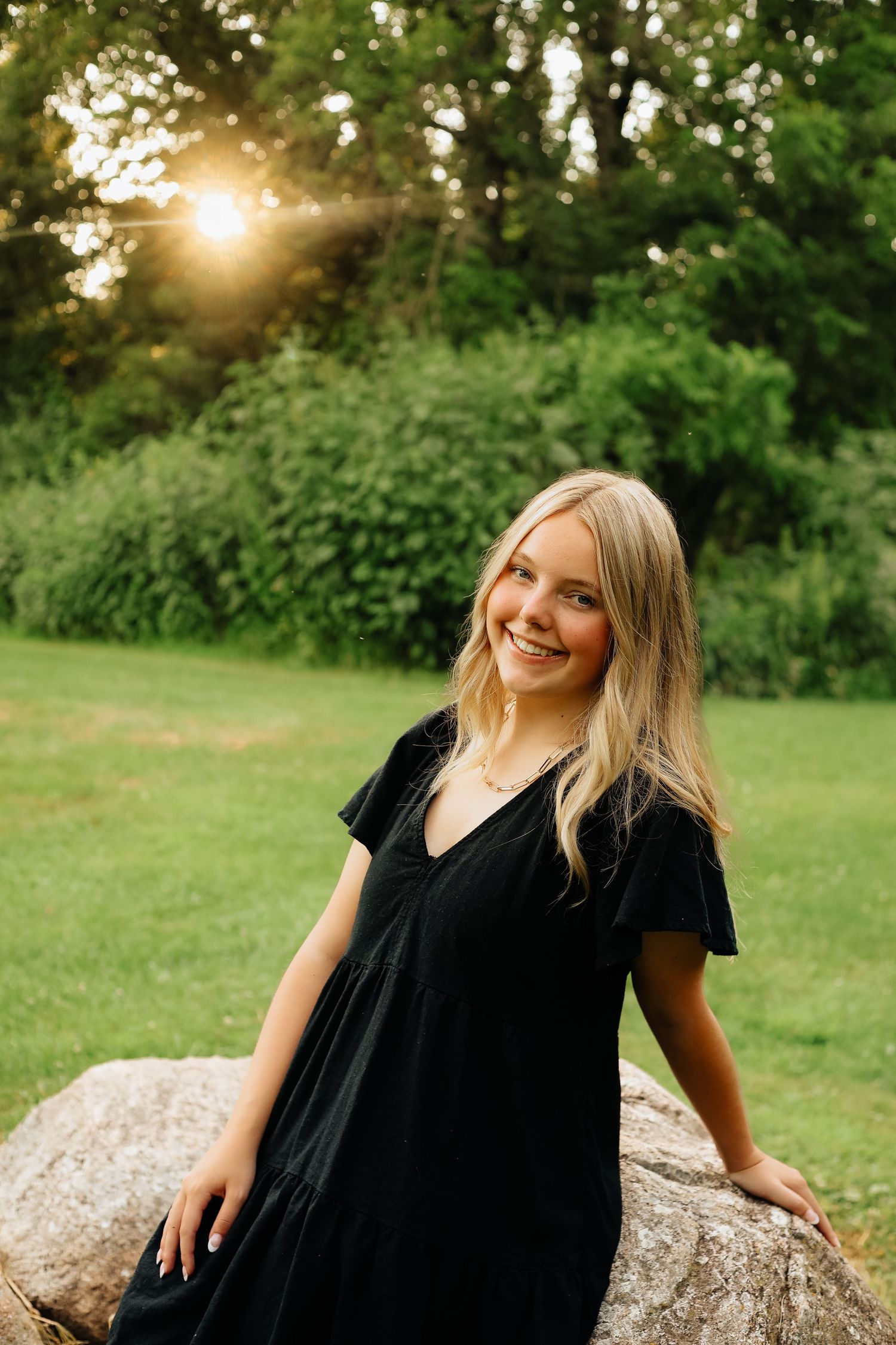 A woman in black dress leans against a large rock in a sunlit garden setting.