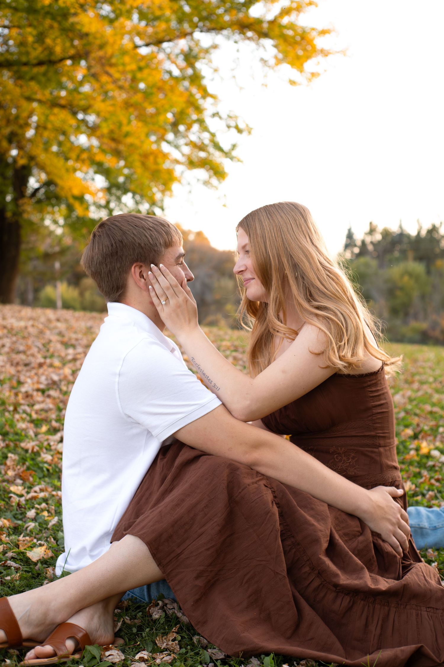 Couple in a brown dress and white shirt sit on autumn leaves, gently touching faces beneath a golden tree at sunset.
