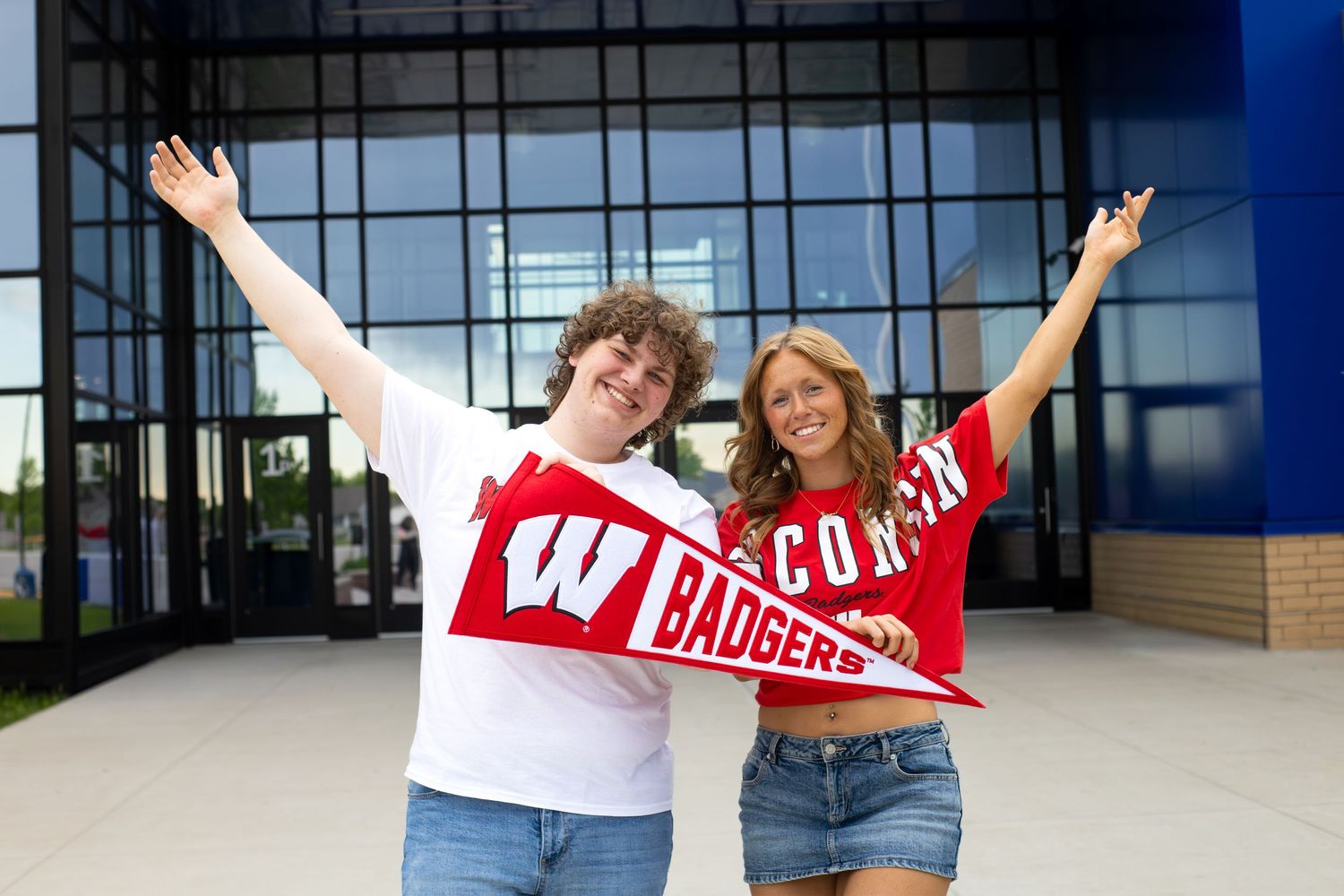 Two Wisconsin Badgers fans celebrate outdoors, holding red pennants and scarves with arms raised joyfully.