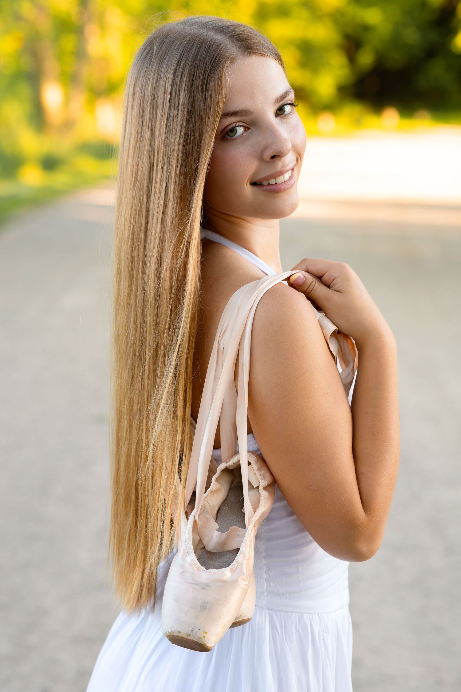 Young blonde ballerina in white dress holding pointe shoes over shoulder, smiling outdoors on a sunlit path.