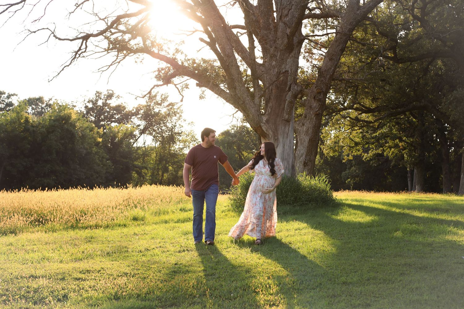 Silhouetted couple walking through sunlit field with large trees in background.